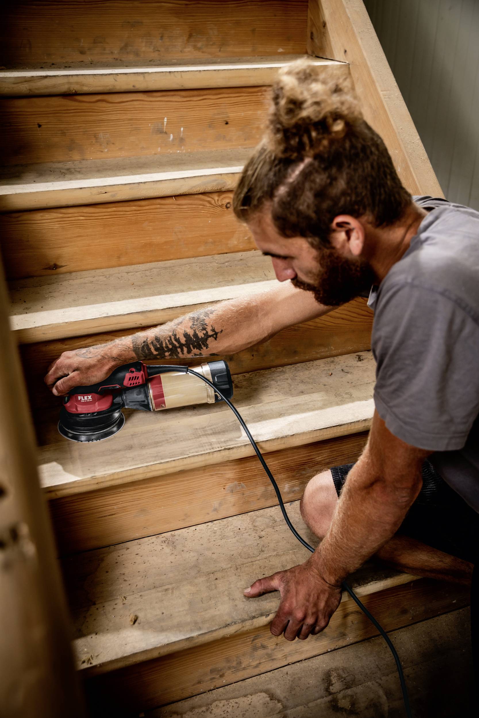Eine Person schleift eine hölzerne Treppe mit einer elektrischen Schleifmaschine. Sie hat ein Tattoo am Arm und trägt ein graues T-Shirt.