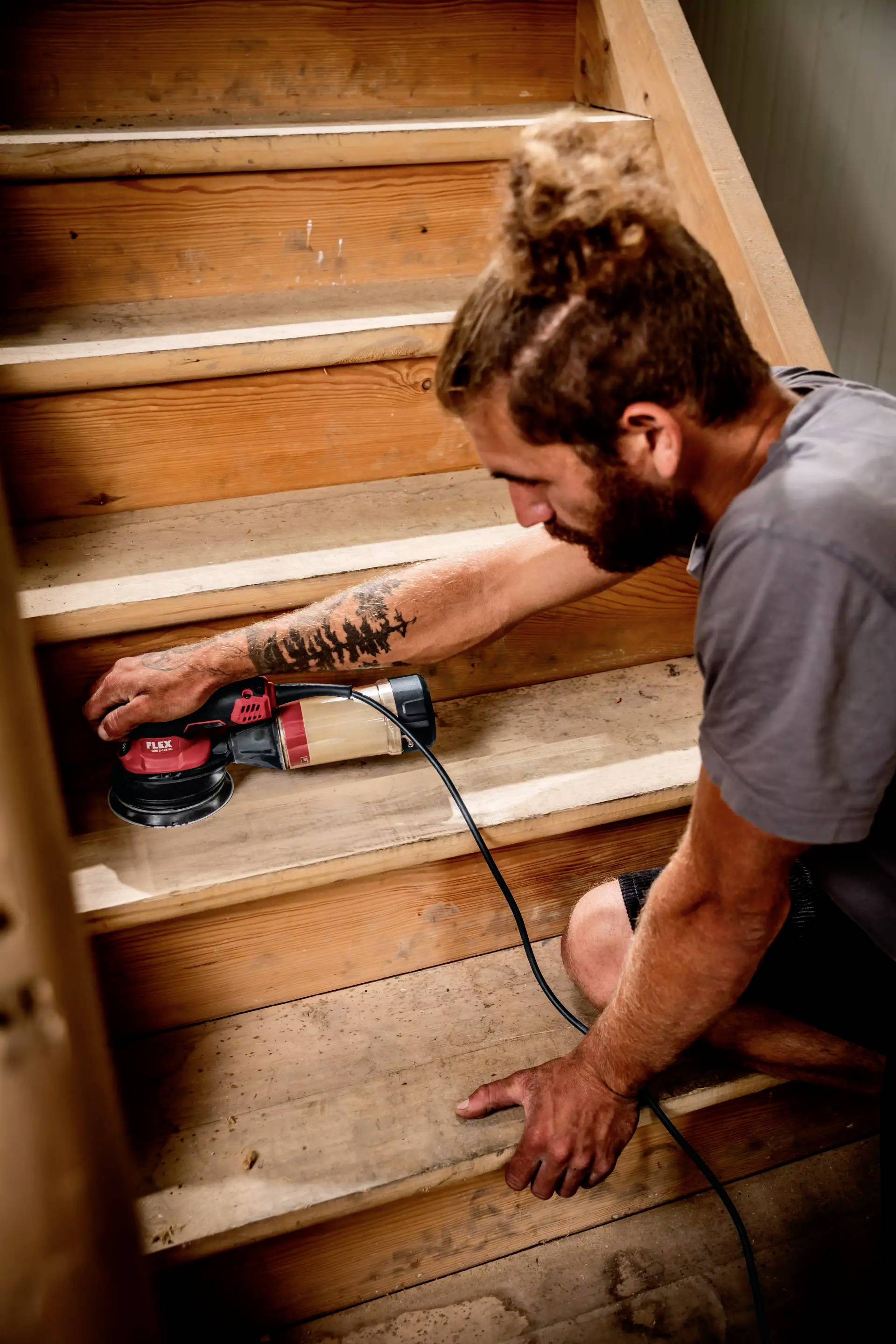 Ein Mann schleift mit einer elektrischen Schleifmaschine eine Holztreppe ab. Er trägt lässige Kleidung und konzentriert sich auf seine Arbeit.