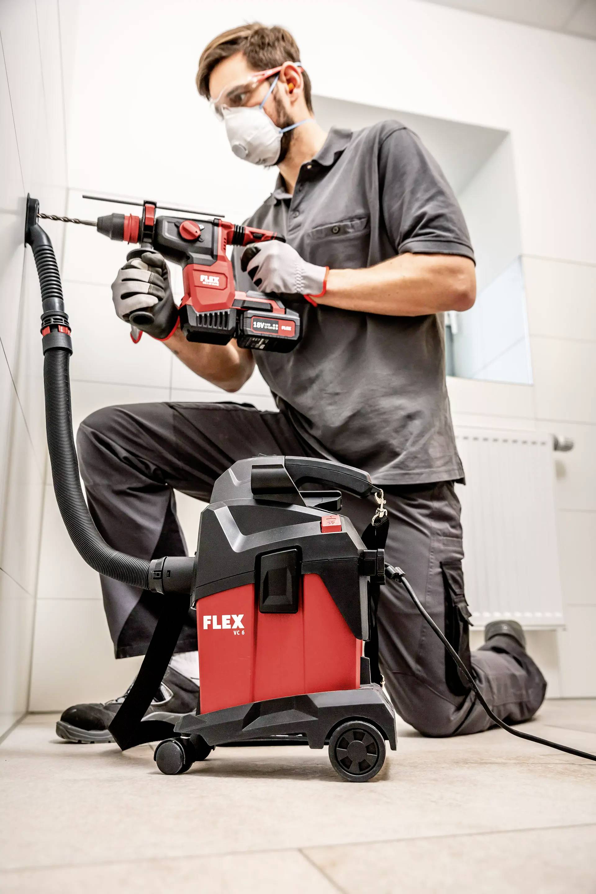Ein Mann mit Schutzbrille und Maske bohrt mit einem roten Bohrer in eine Wand. Vor ihm steht ein Staubsauger, der Staub absaugt.