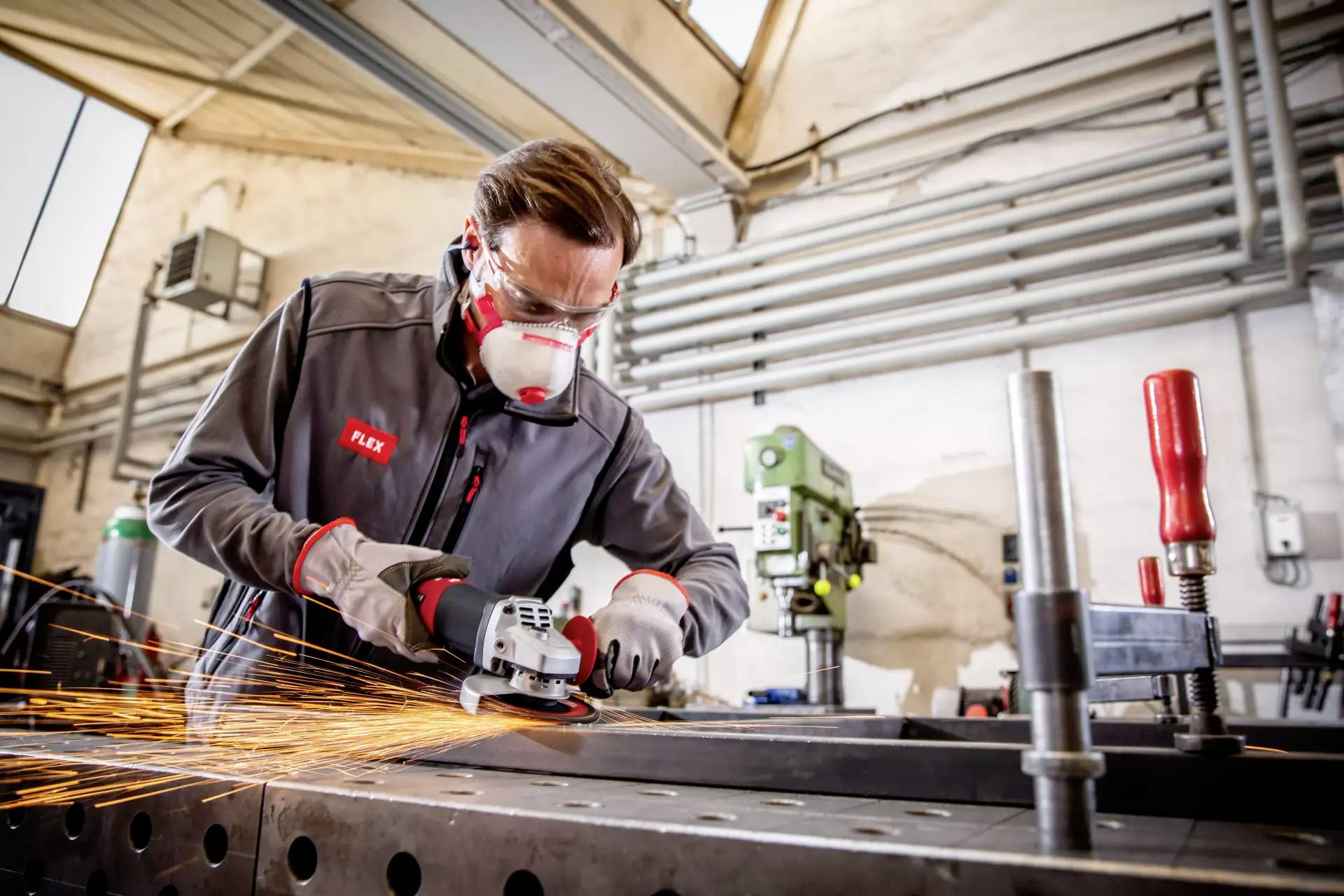 Eine Person in Schutzkleidung schleift Metall mit einem Winkelschleifer. Funken fliegen, während sie in einer Werkstatt arbeitet.