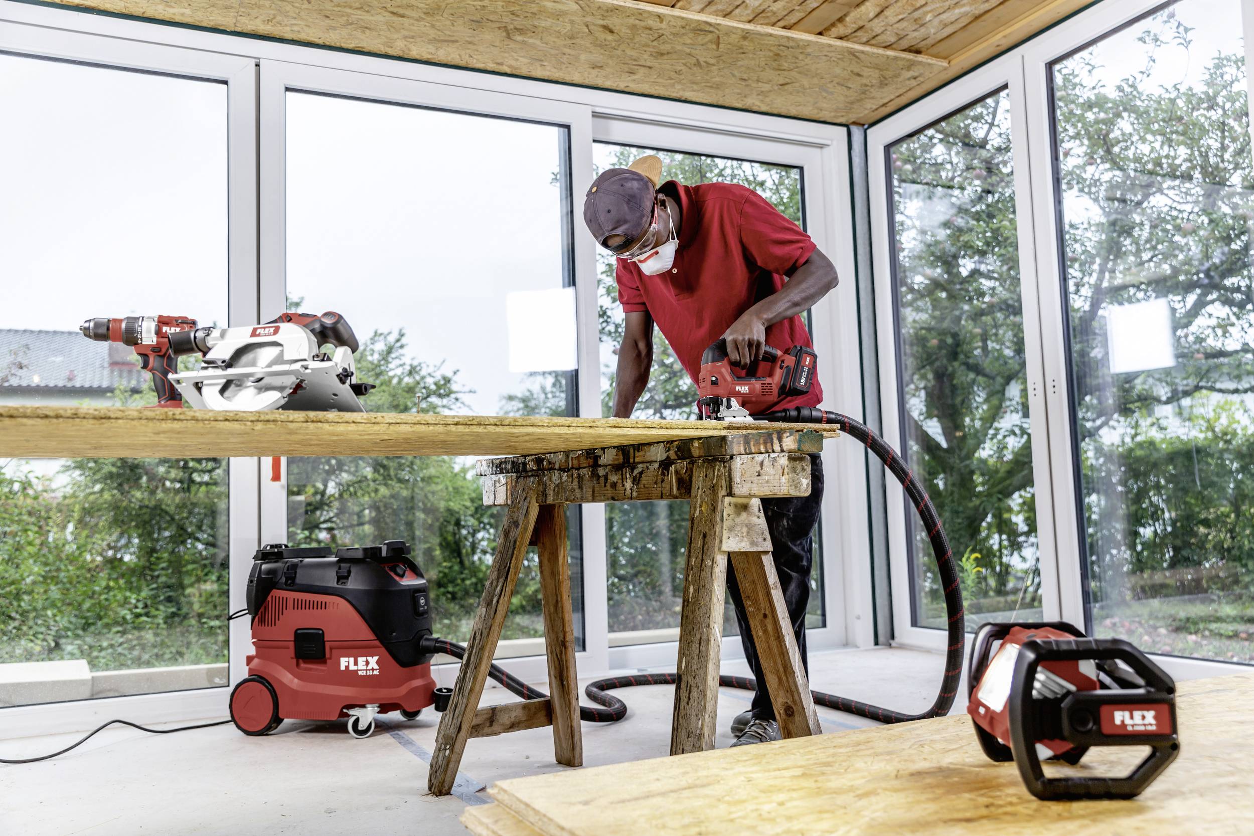 Ein Handwerker bearbeitet Holz in einem lichtdurchfluteten Raum, umgeben von Werkzeugen. Draußen sind Bäume sichtbar.
