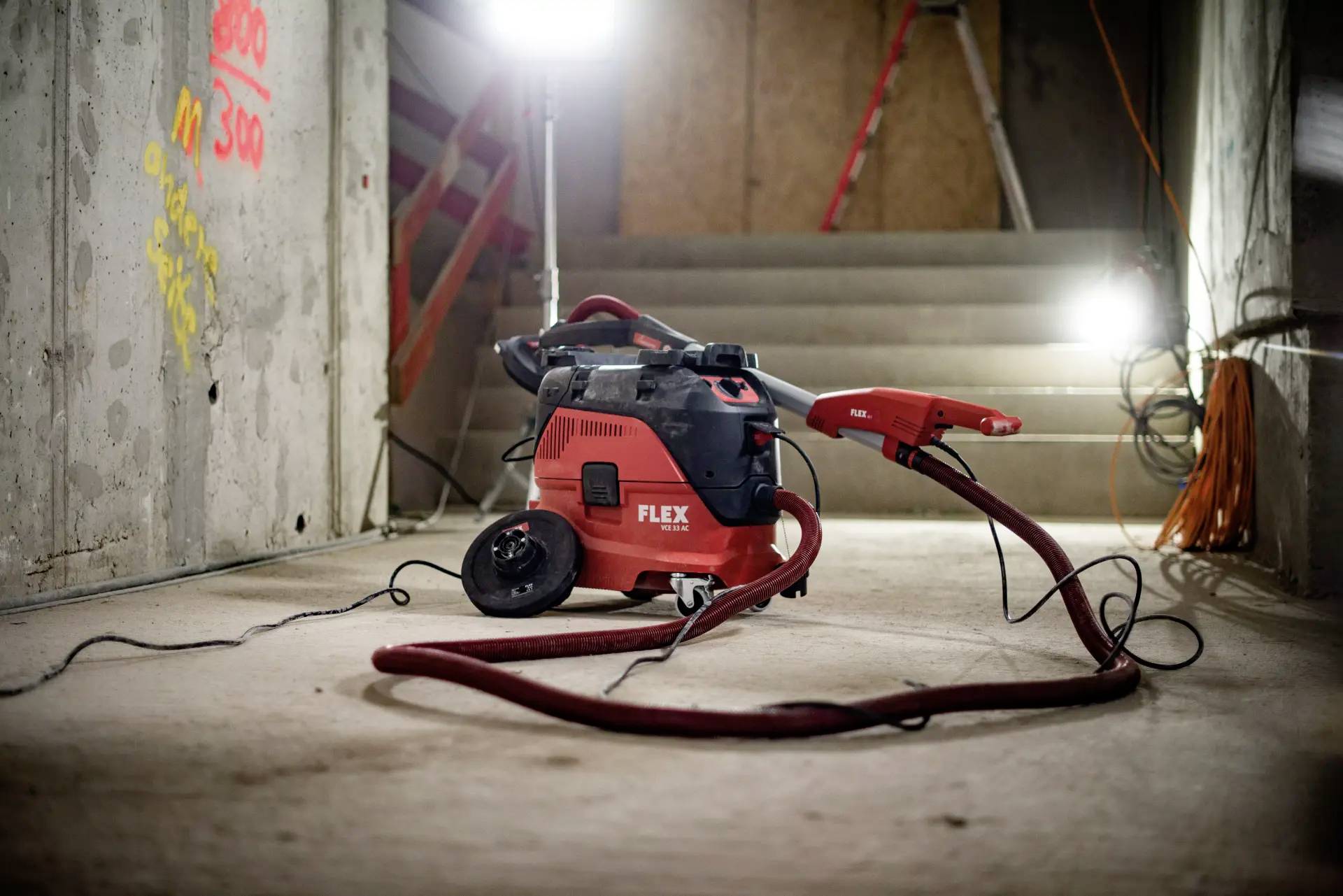 Ein roter Industriesauger steht in einem schwach beleuchteten Kellerraum, von Baustrahlern beleuchtet, neben baustellentypischen Kabeln.