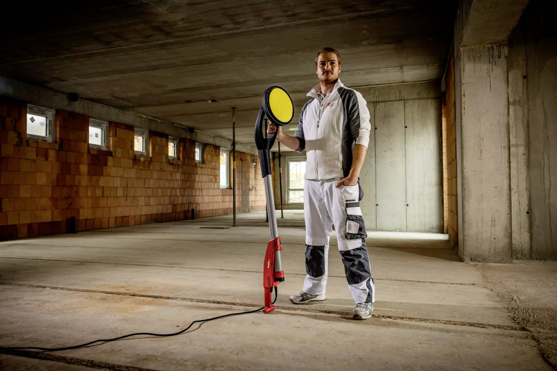 Ein Mann in Arbeitskleidung steht in einem unvollständigen Betonraum und hält ein großes Elektrogerät. Die Umgebung wirkt industriell.