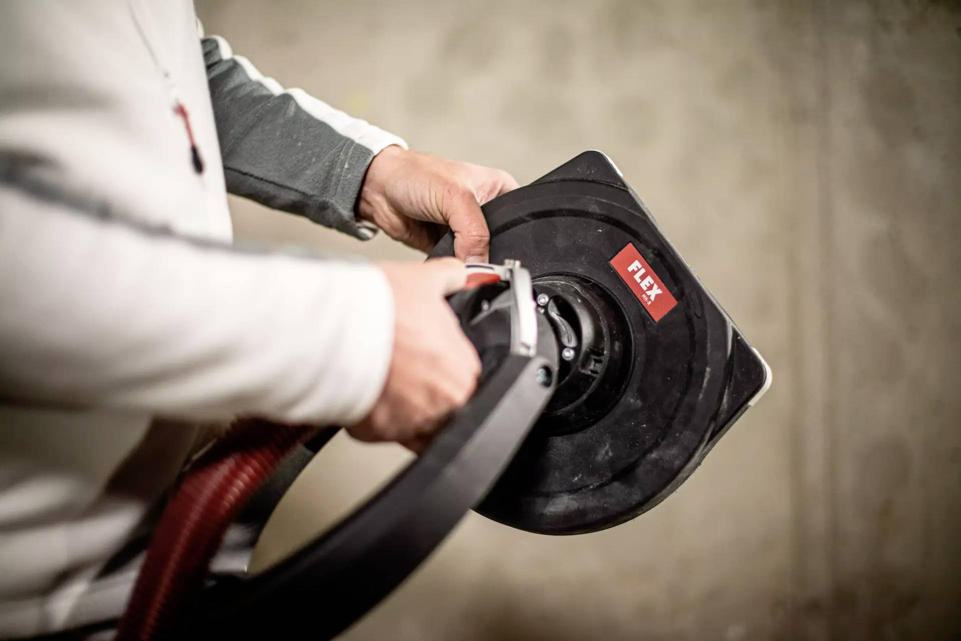 Eine Person hält ein elektrisches Schleifgerät mit rotem Schlauch, der Flex-Marke. Das Gerät wird offensichtlich in einer Werkstatt verwendet.