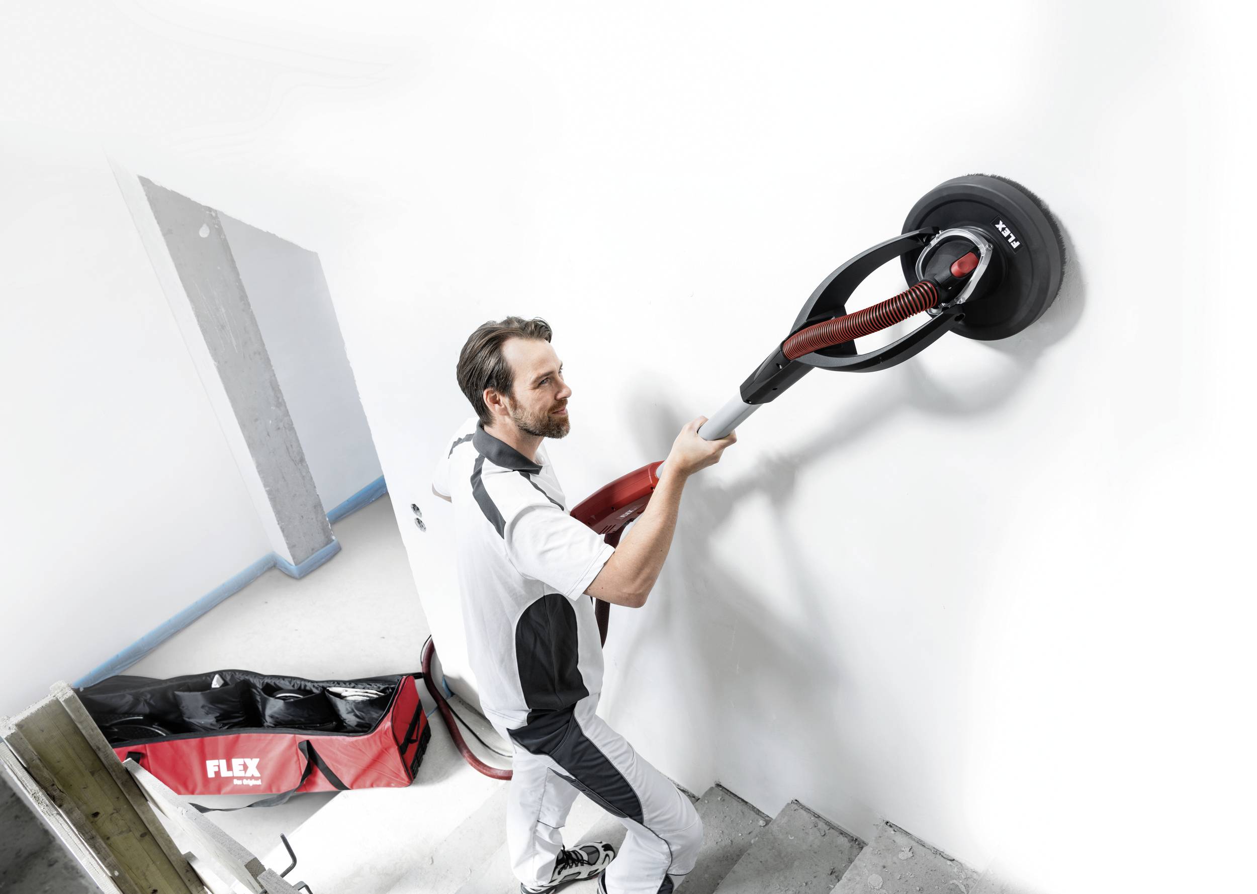 Ein Mann schleift mit einer elektrischen Schleifmaschine eine weiße Wand. Neben ihm steht eine rote Tasche mit der Aufschrift 'FLEX'.