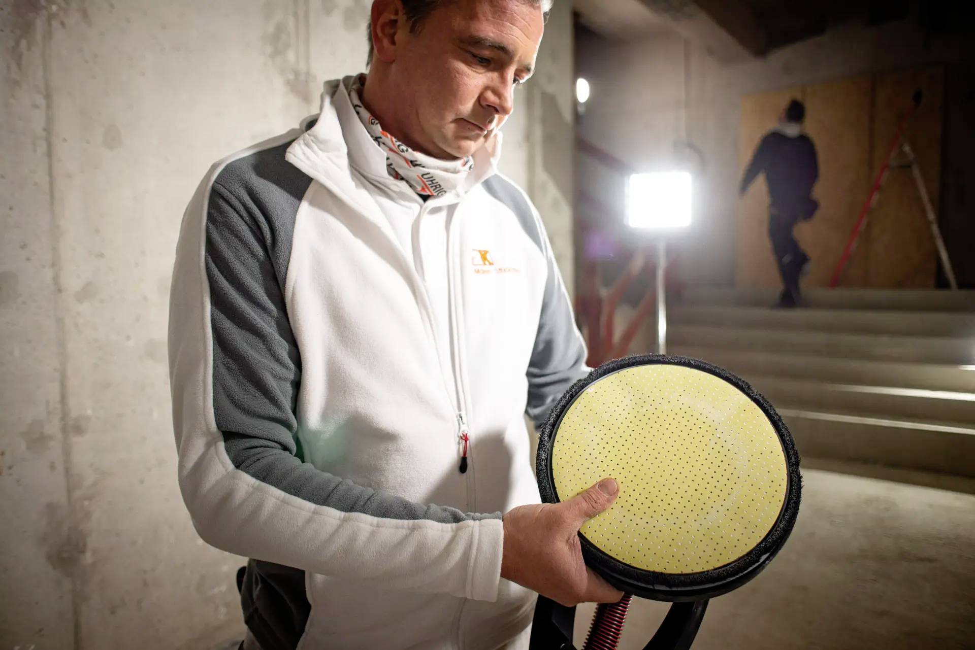 Ein Mann in Arbeitskleidung steht in einem Keller und hält eine runde, gelbe Schleifscheibe, während im Hintergrund eine Person vorbeigeht.