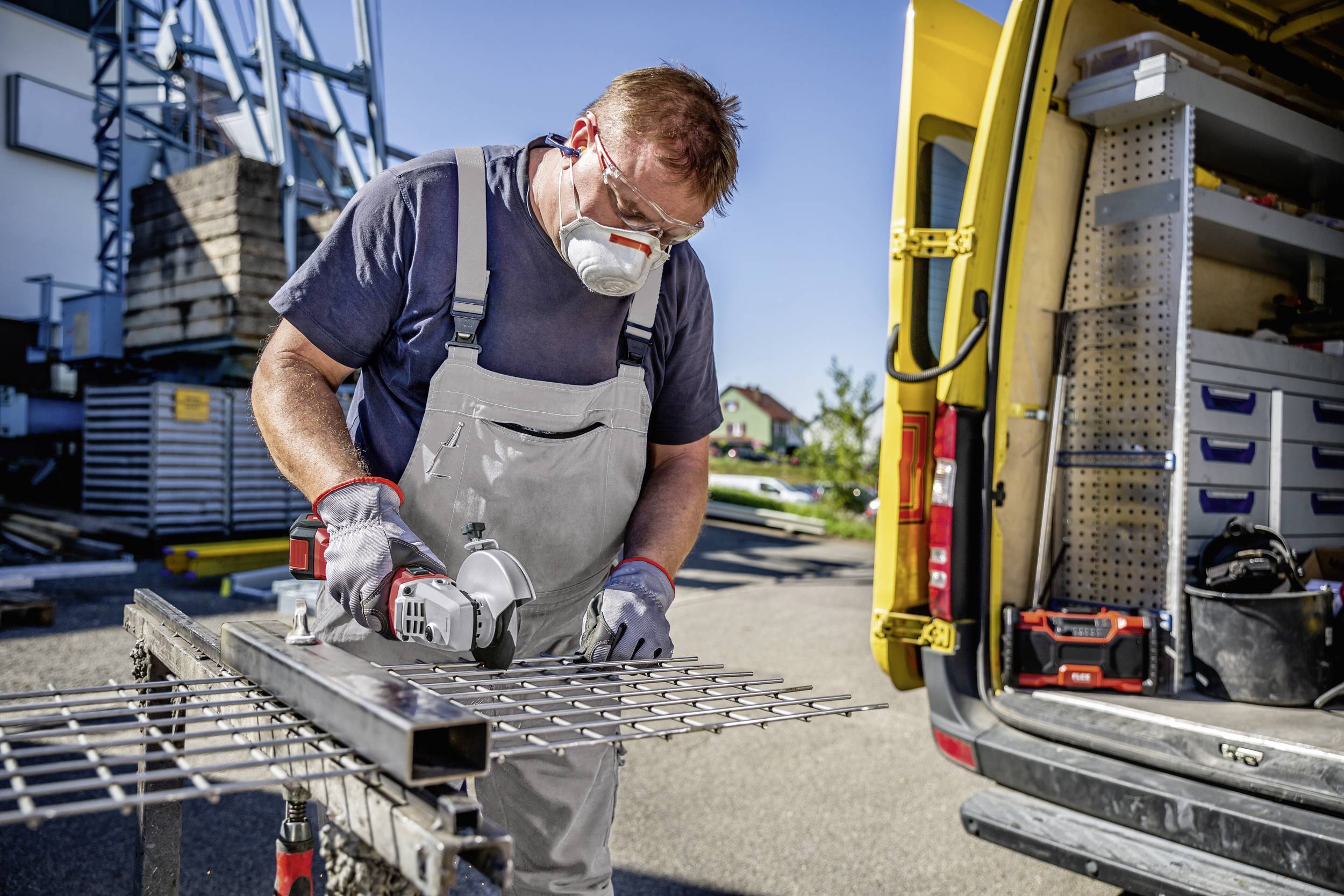 Ein Mann in Arbeitskleidung und Schutzbrille schneidet ein Metallgitter mit einer Trennscheibe neben einem gelben Handwerkerfahrzeug.
