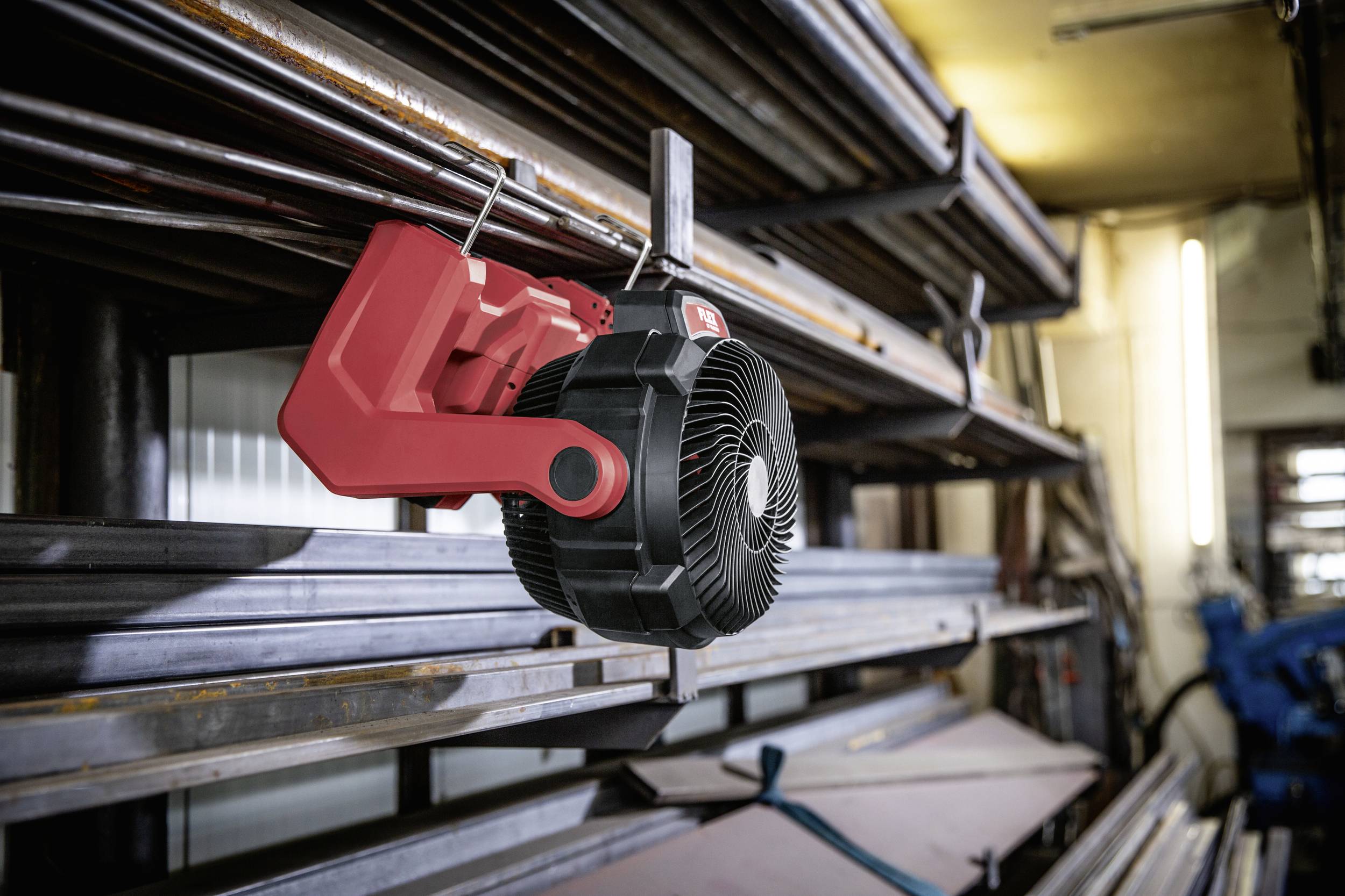 Ein roter Akku-Ventilator hängt an einer Metallstange in einer Werkstatt. Im Hintergrund sind gestapelte Metallbalken sichtbar.