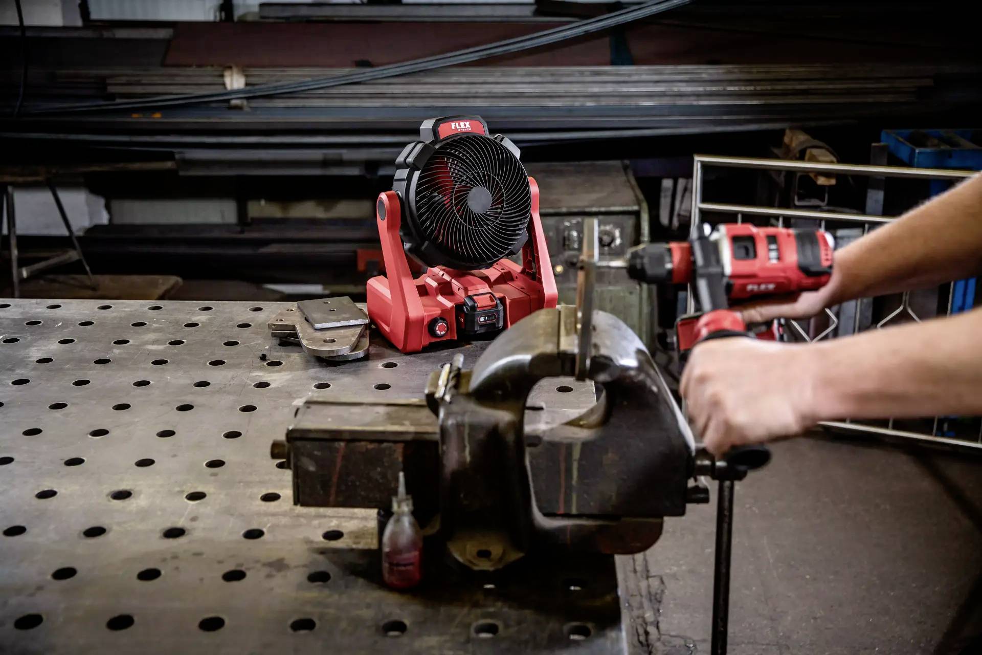 Eine Werkbank mit einem Schraubstock und einer Bohrmaschine in Gebrauch; im Hintergrund steht ein tragbarer Ventilator auf einem Tisch.