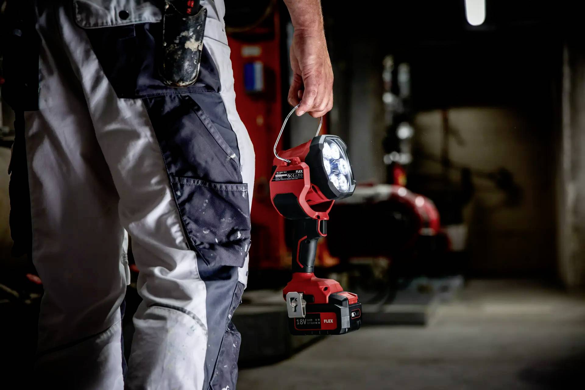 Ein Arbeiter in Arbeitskleidung hält eine tragbare LED-Lampe in einem düsteren, industriellen Keller. Im Hintergrund sind große Maschinen sichtbar.