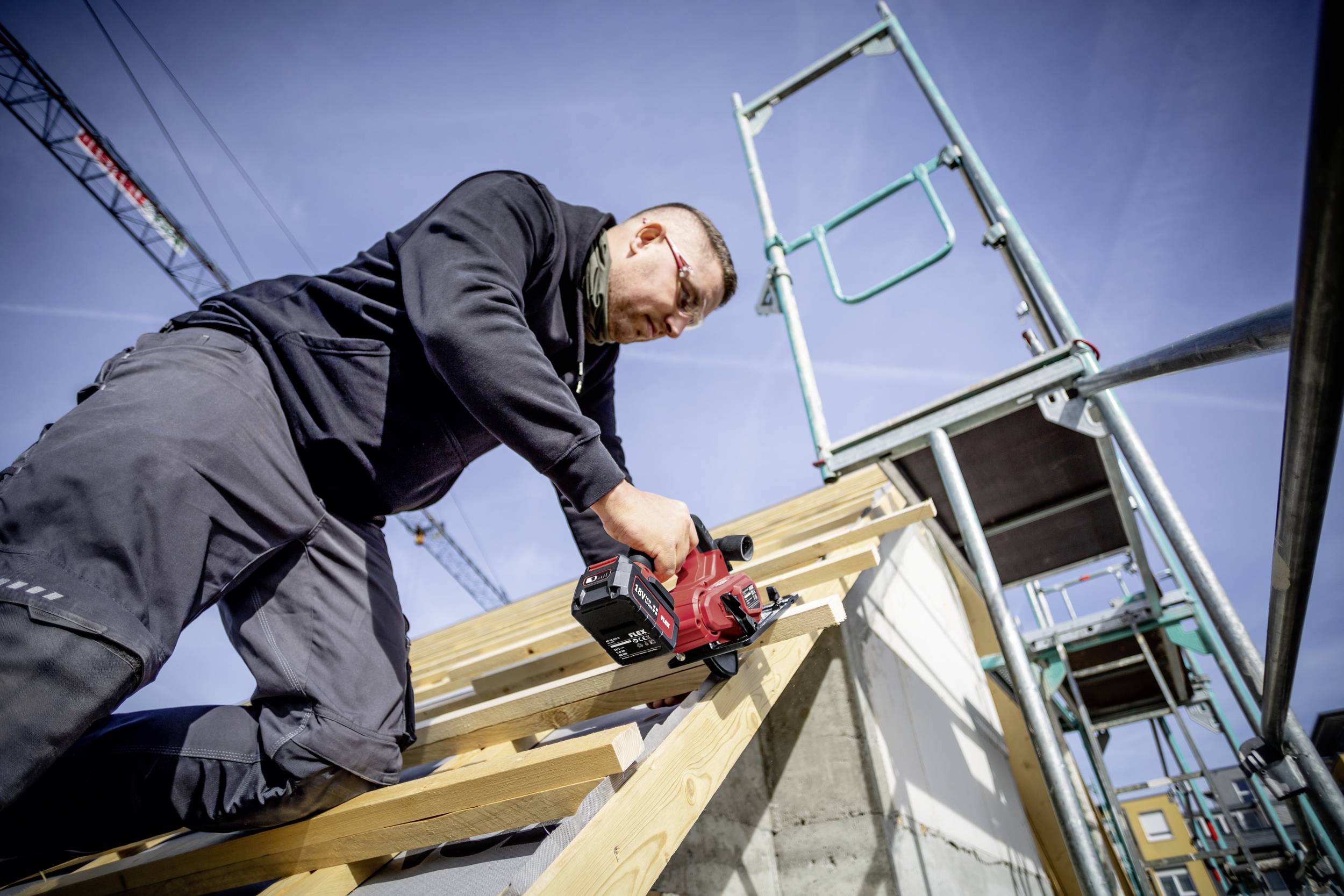 Ein Arbeiter auf einer Baustelle verwendet ein Elektrowerkzeug, um Holzlatten zu befestigen. Im Hintergrund sind Gerüste zu sehen.
