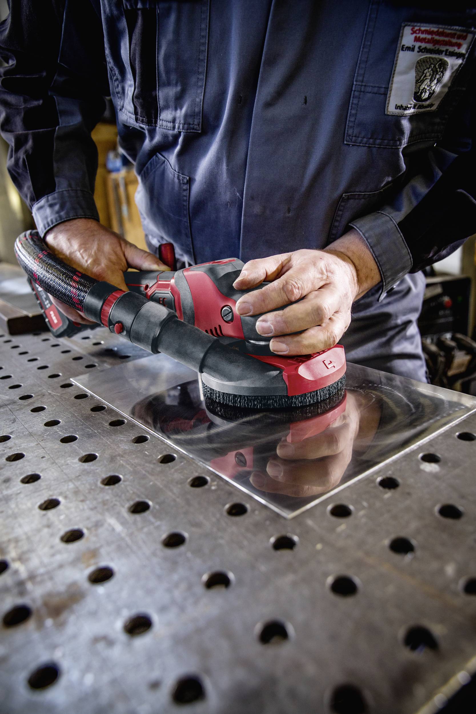 Person schleift Metallplatte mit einer roten Schleifmaschine. Trägt blaue Arbeitskleidung, steht an werkzeugübersätem Arbeitstisch.