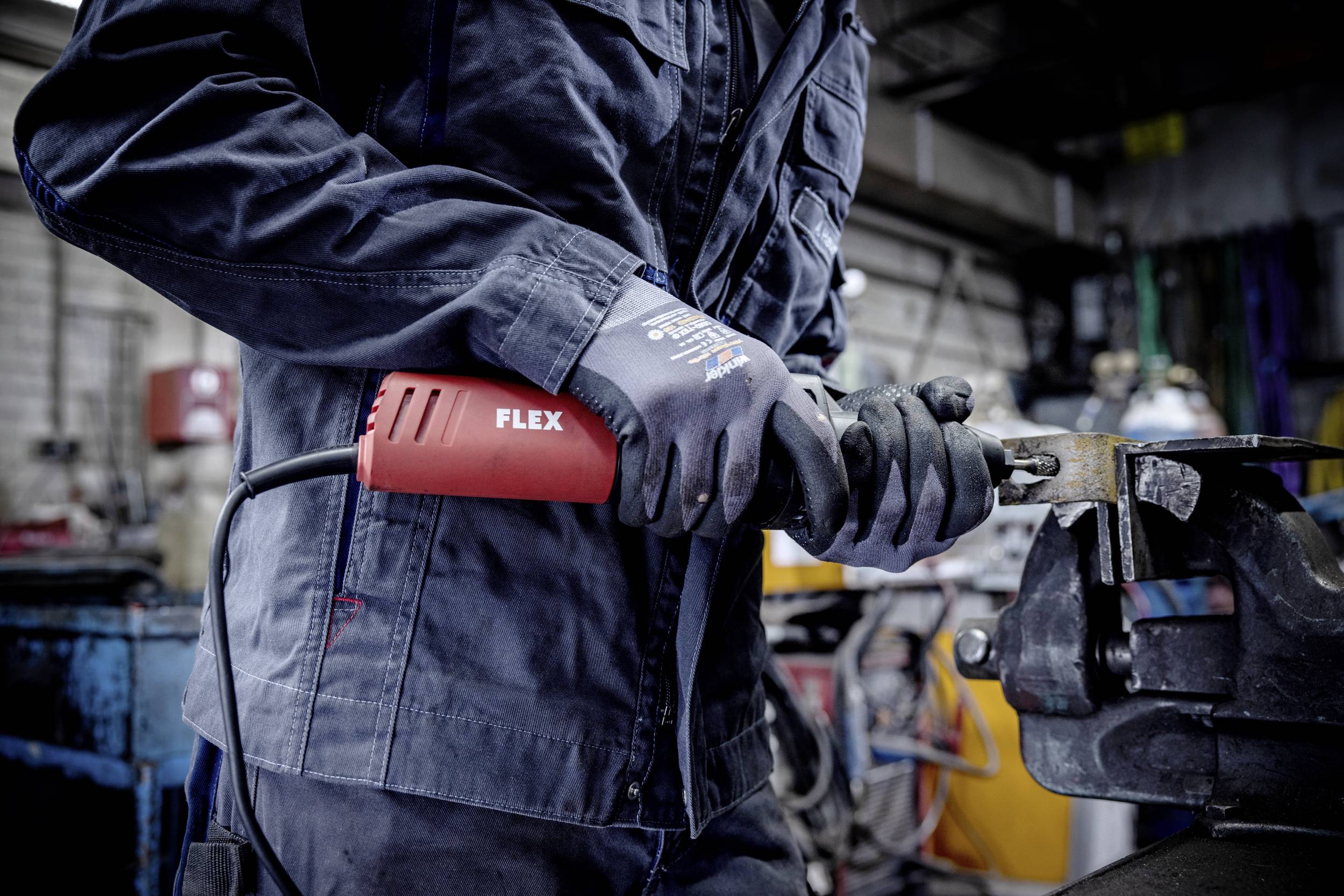 Eine Person in Arbeitskleidung hält ein elektrisches Werkzeug in der Hand, vermutlich eine Schleifmaschine, in einer Werkstattumgebung.