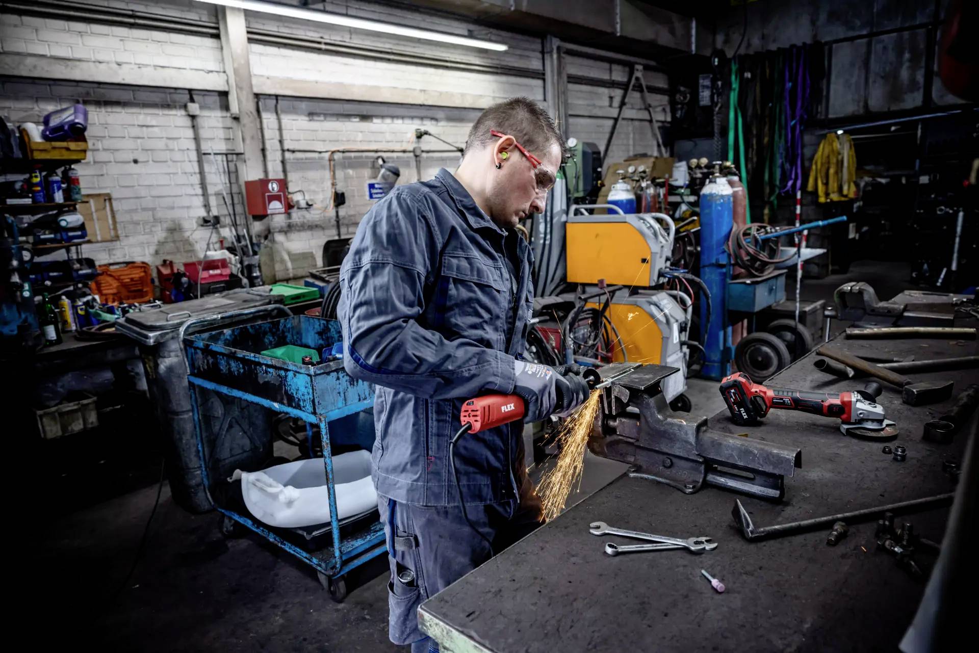 Ein Mann schleift Metall in einer Werkstatt, Funken sprühen. Im Hintergrund sind Werkzeuge und Maschinen. Er trägt Schutzbrille.