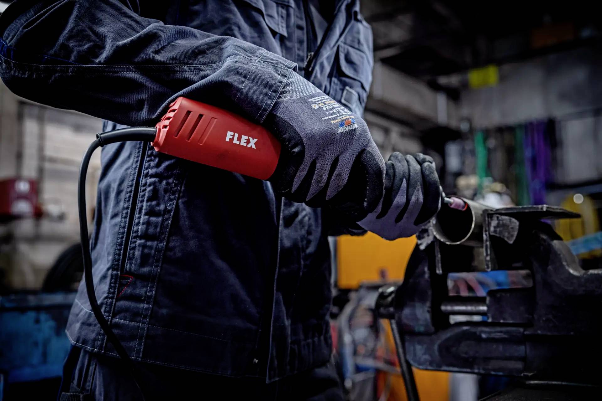 Person in Arbeitskleidung benutzt eine elektrische Flex-Schleifmaschine, um ein Metallrohr in einer Werkstattumgebung zu bearbeiten.