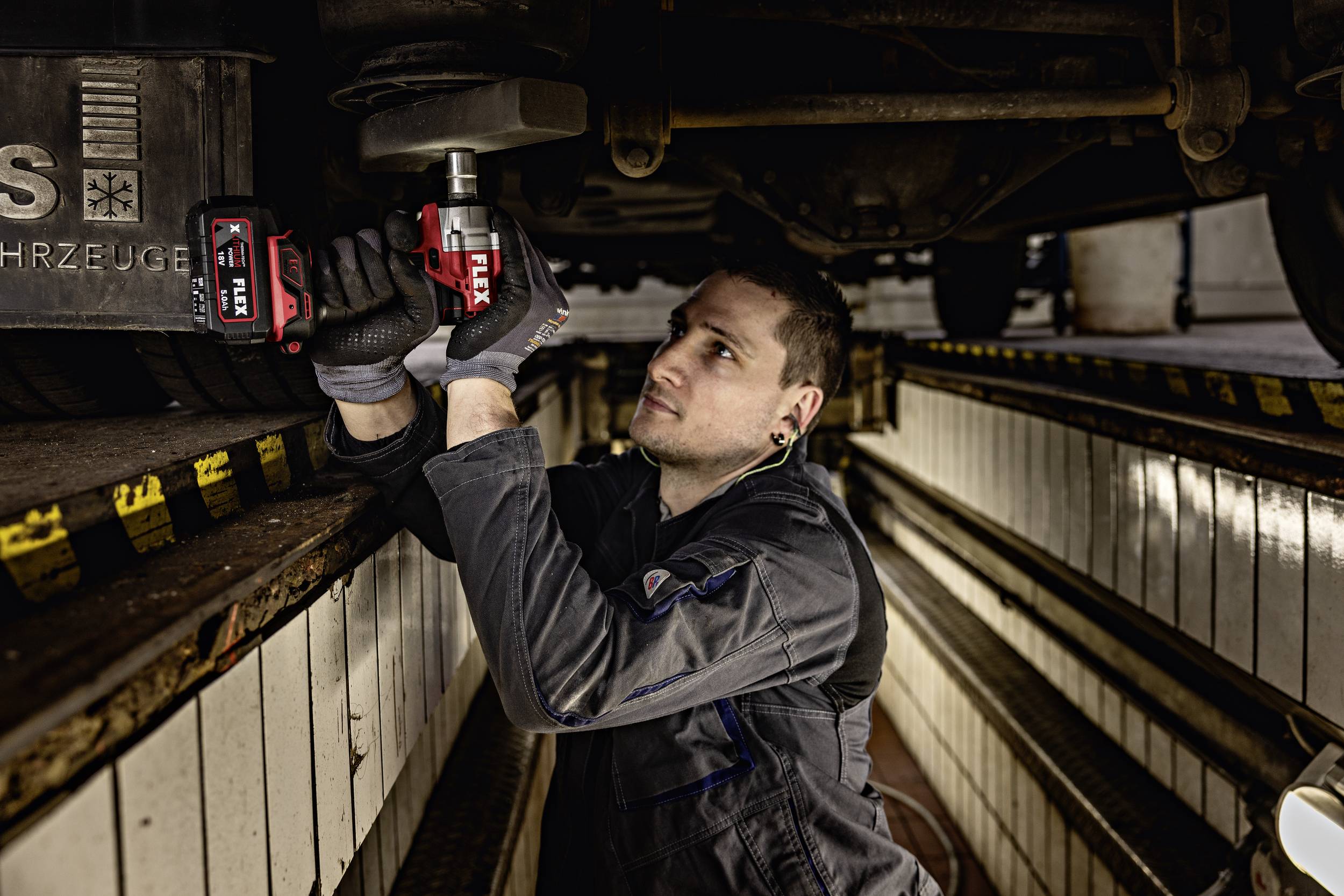 Ein Mechaniker liegt in einer Werkstattgrube und repariert die Unterseite eines Fahrzeugs mit einem Elektrowerkzeug.