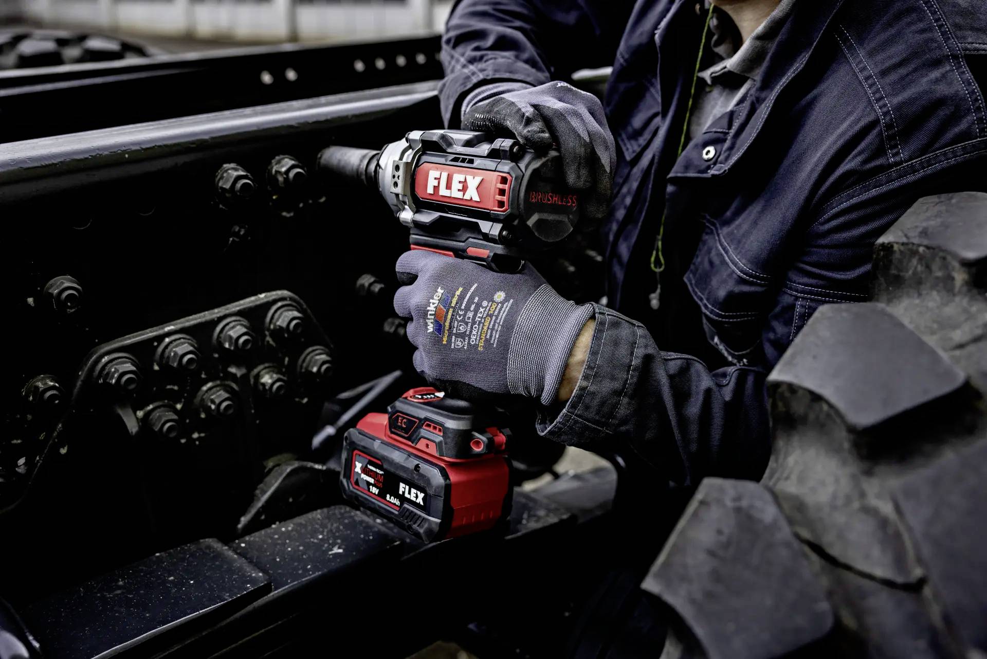 Person hält eine rote elektrische Bohrmaschine an einem Fahrzeugrahmen fest. Werkzeug ist als 'FLEX' gekennzeichnet. Fokus auf Arbeit.
