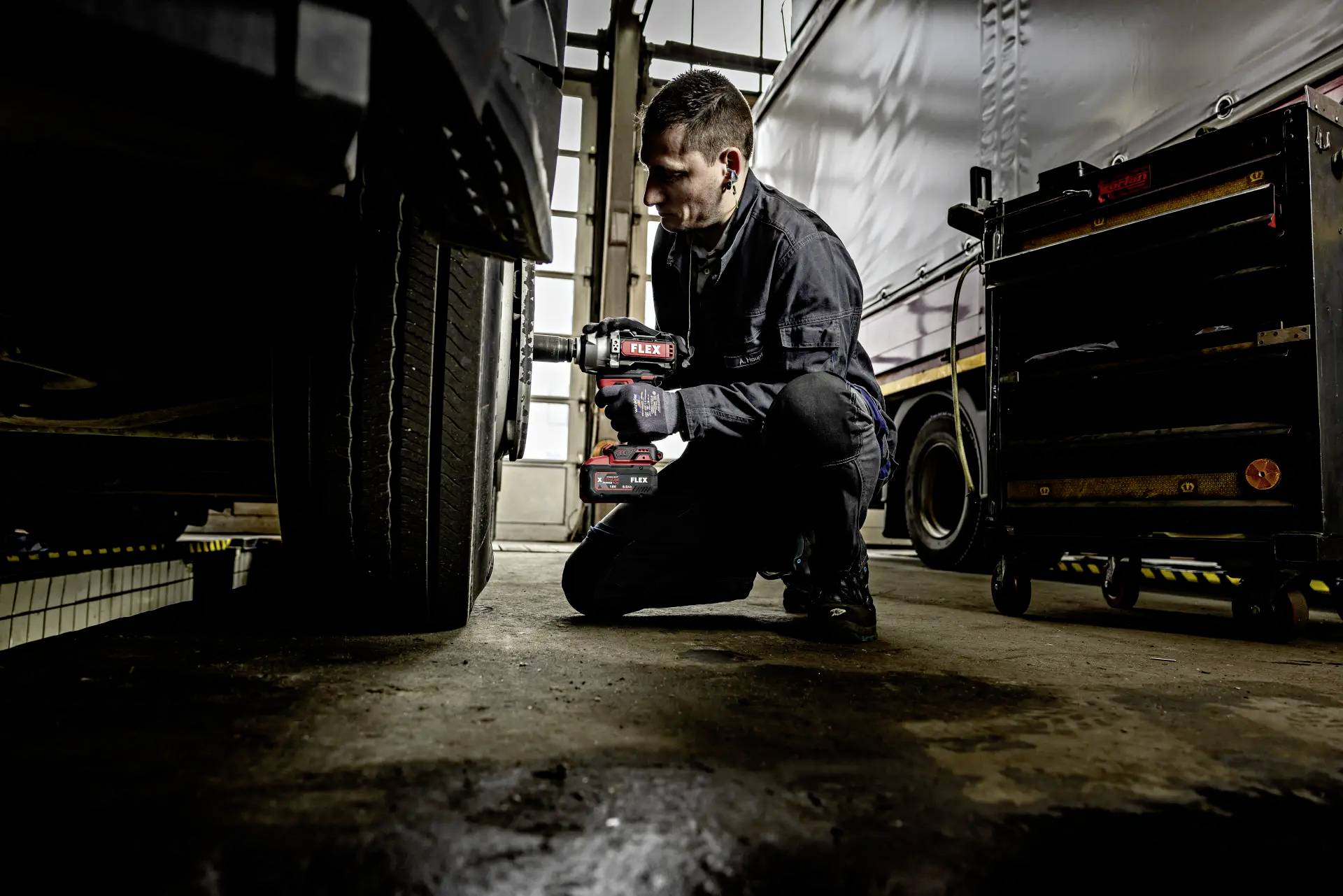 Mechaniker überprüft Autoreifen mit einem Elektrowerkzeug in einer Werkstatt; Fahrzeug und Werkzeugschrank im Hintergrund.