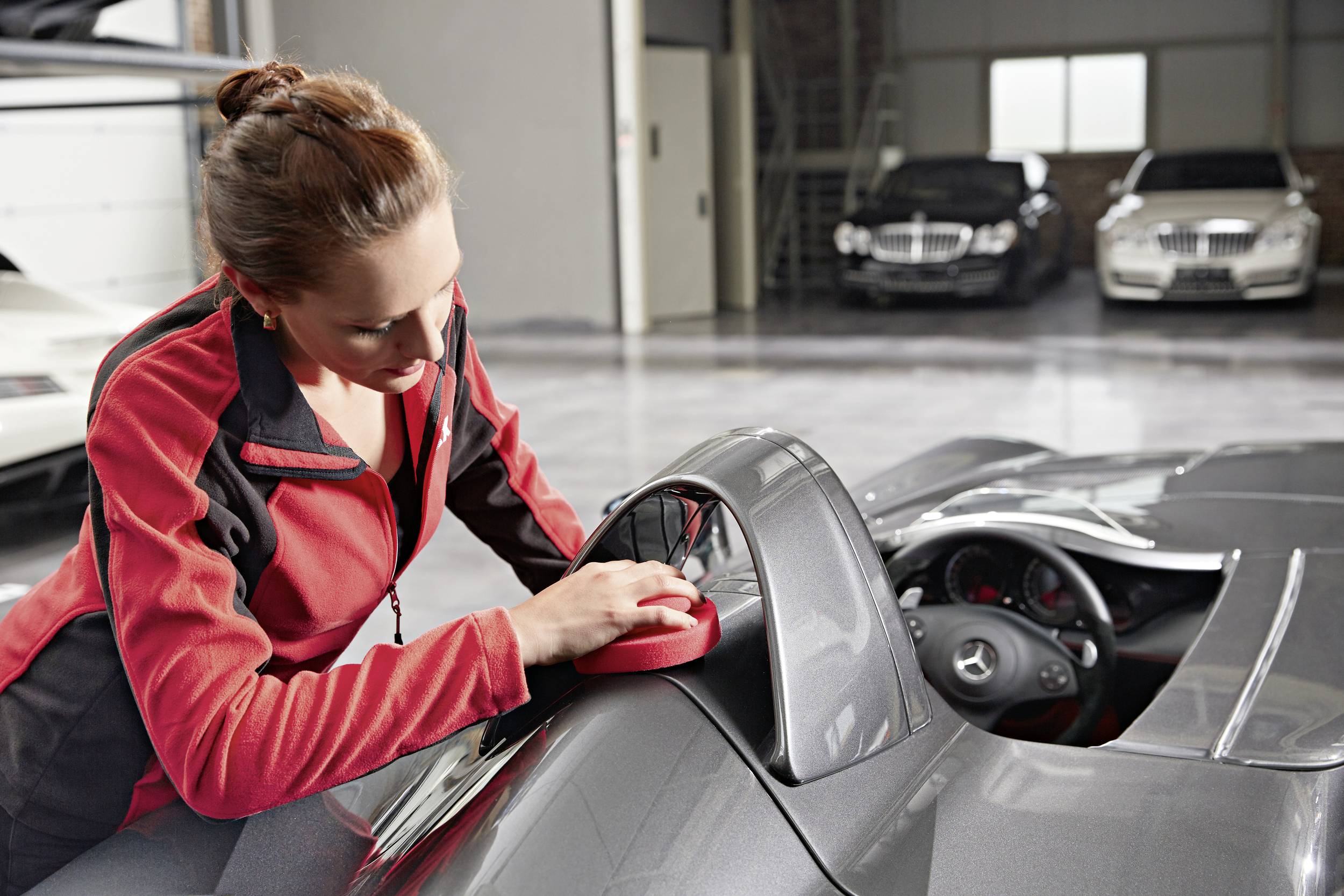 Eine Frau poliert einen grauen Sportwagen in einer Autowerkstatt. Im Hintergrund stehen andere Luxusfahrzeuge.