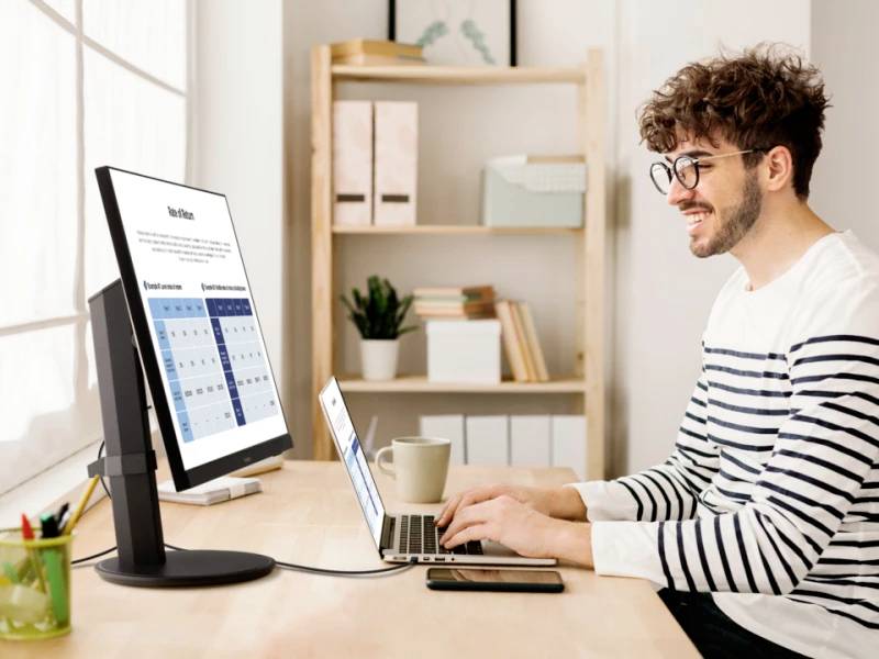 Ein Mann arbeitet lächelnd am Laptop an einem Schreibtisch, mit einem Monitor und einer Tasse Kaffee. Regale im Hintergrund.