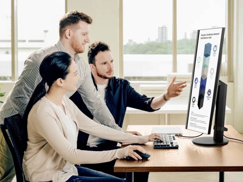 Drei Personen betrachten zusammen einen großen Computermonitor mit einer Infografik. Ein Mann zeigt auf den Bildschirm, während sie diskutieren.