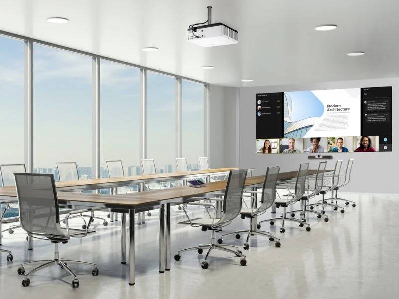 Großer Konferenzraum mit rechteckigem Tisch und mehreren Bürostühlen. An der Wand ein großer Bildschirm mit einer Videokonferenz.
