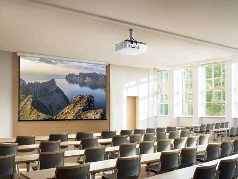 Ein leerer Klassenraum mit Projektion einer Landschaft mit Bergen und Meer auf der Leinwand. Große Fenster lassen Tageslicht herein.