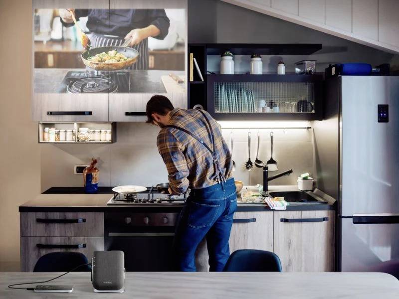 Ein Mann kocht in einer modernen Küche, während ein Kochvideo auf einem Bildschirm gezeigt wird. Er trägt ein kariertes Hemd und Hosenträger.