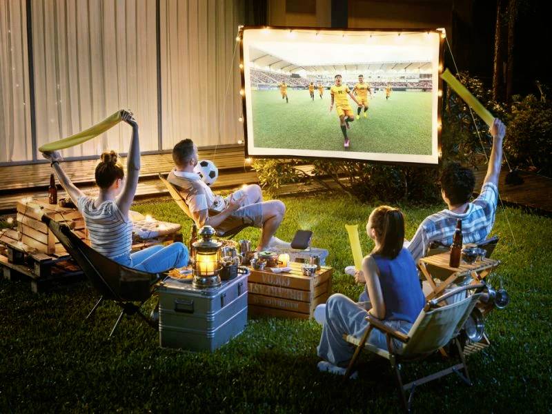 Vier Personen sitzen im Garten und schauen ein Fußballspiel auf eine Leinwand im Freien. Getränke und Snacks stehen bereit.