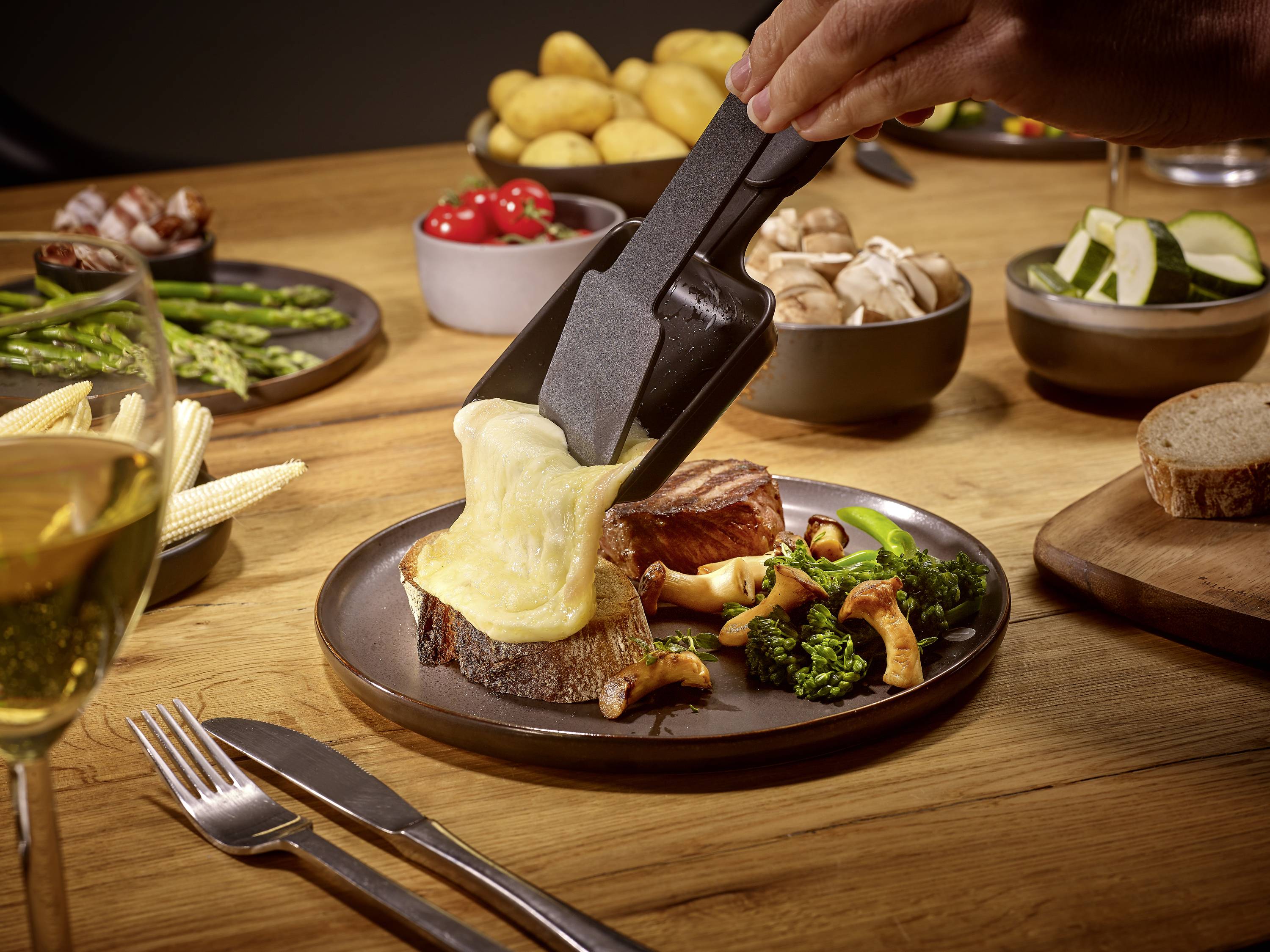 Eine Hand gießt geschmolzenen Käse über ein Stück Brot auf einem Teller mit Gemüse in einem gemütlichen Essenssetting.