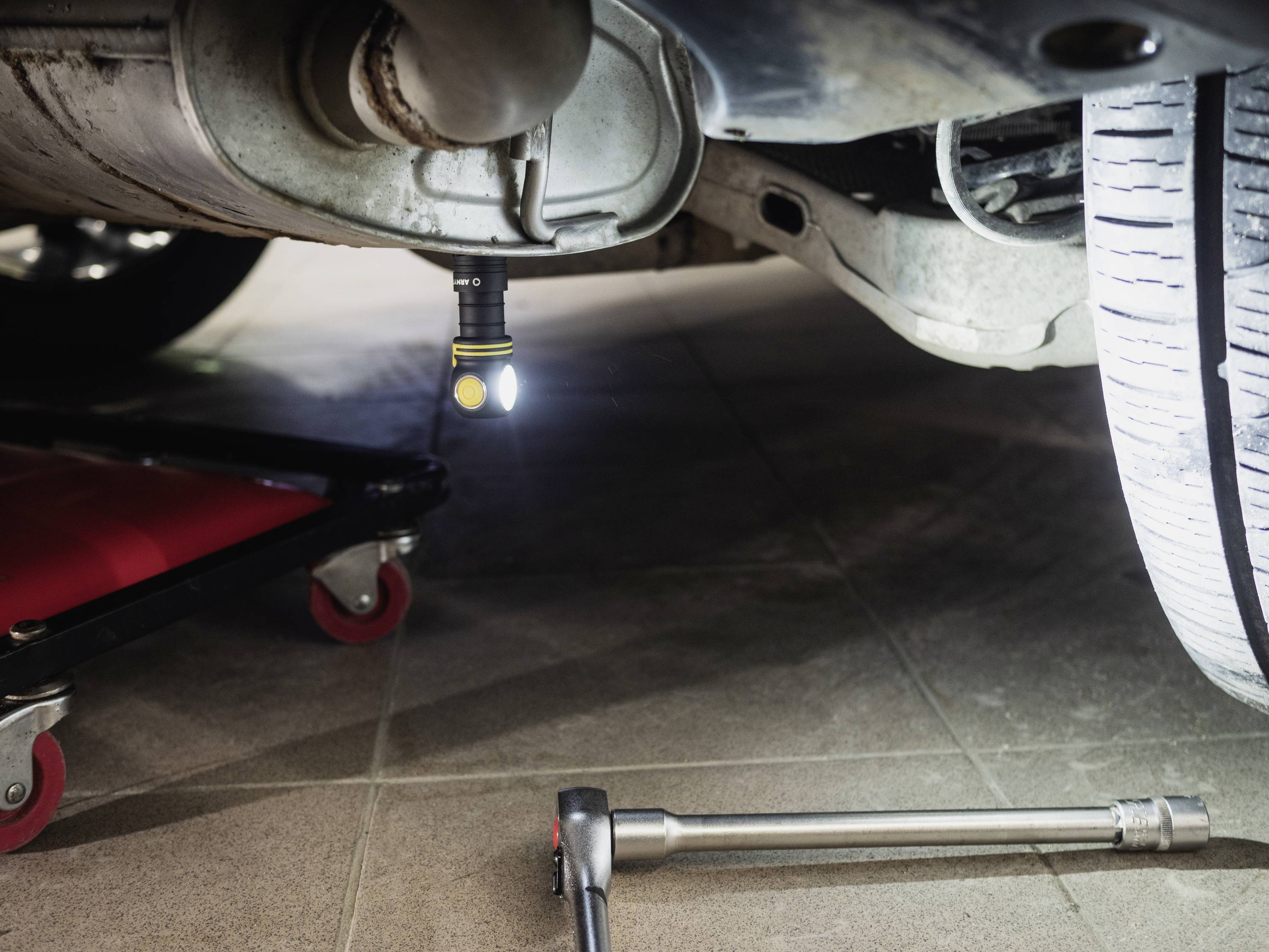 Auto in Werkstatt mit Taschenlampe unter dem Chassis. Eine Hebebühne und Schraubenschlüssel sind sichtbar, Reparatur im Gange.