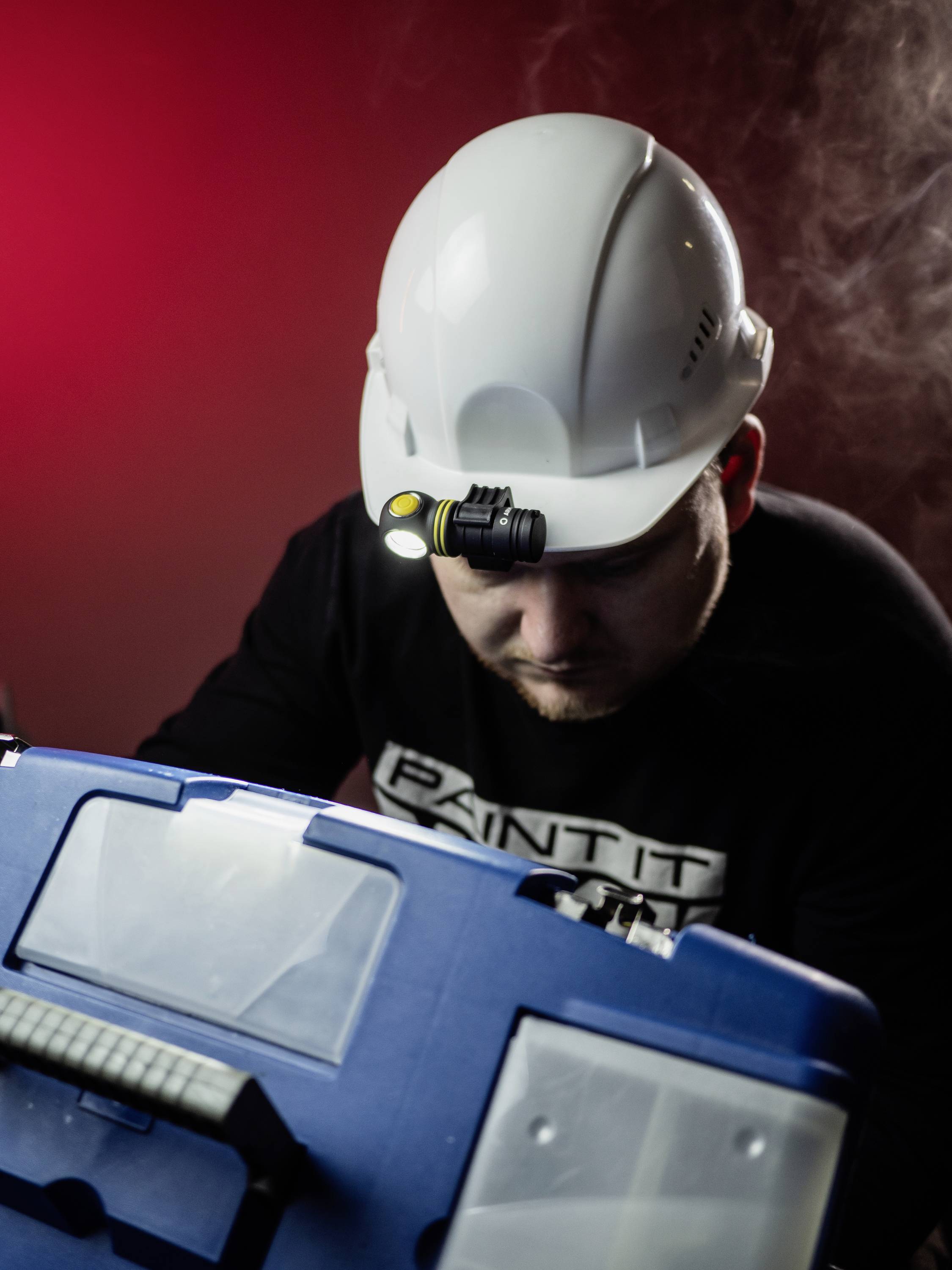 Ein Bauarbeiter mit weißem Schutzhelm und Stirnlampe inspiziert einen blauen Werkzeugkasten in einem schwach beleuchteten Raum.