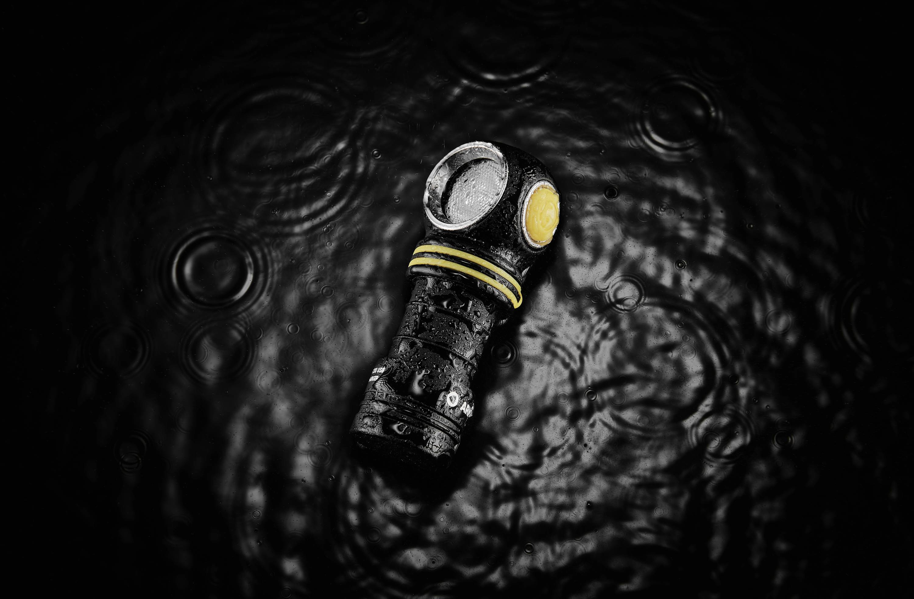 Taschenlampe mit gelbem Knopf in schwarzem Wasser, Wassertropfen darauf, umgeben von konzentrischen Wellen.