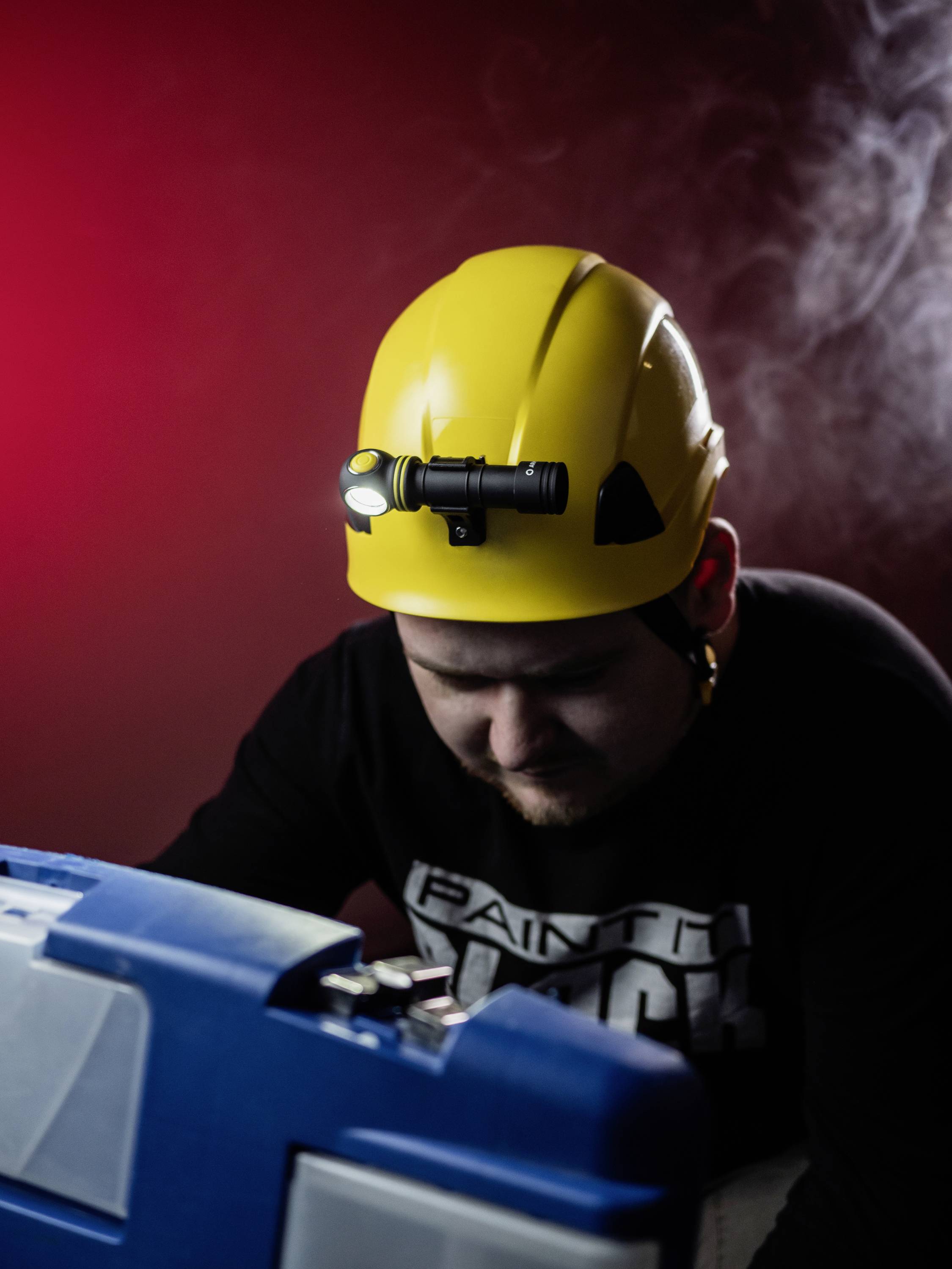Ein Mann trägt einen gelben Schutzhelm mit Stirnlampe, beugt sich über einen blauen Werkzeugkoffer im Licht einer roten Hintergrundbeleuchtung.