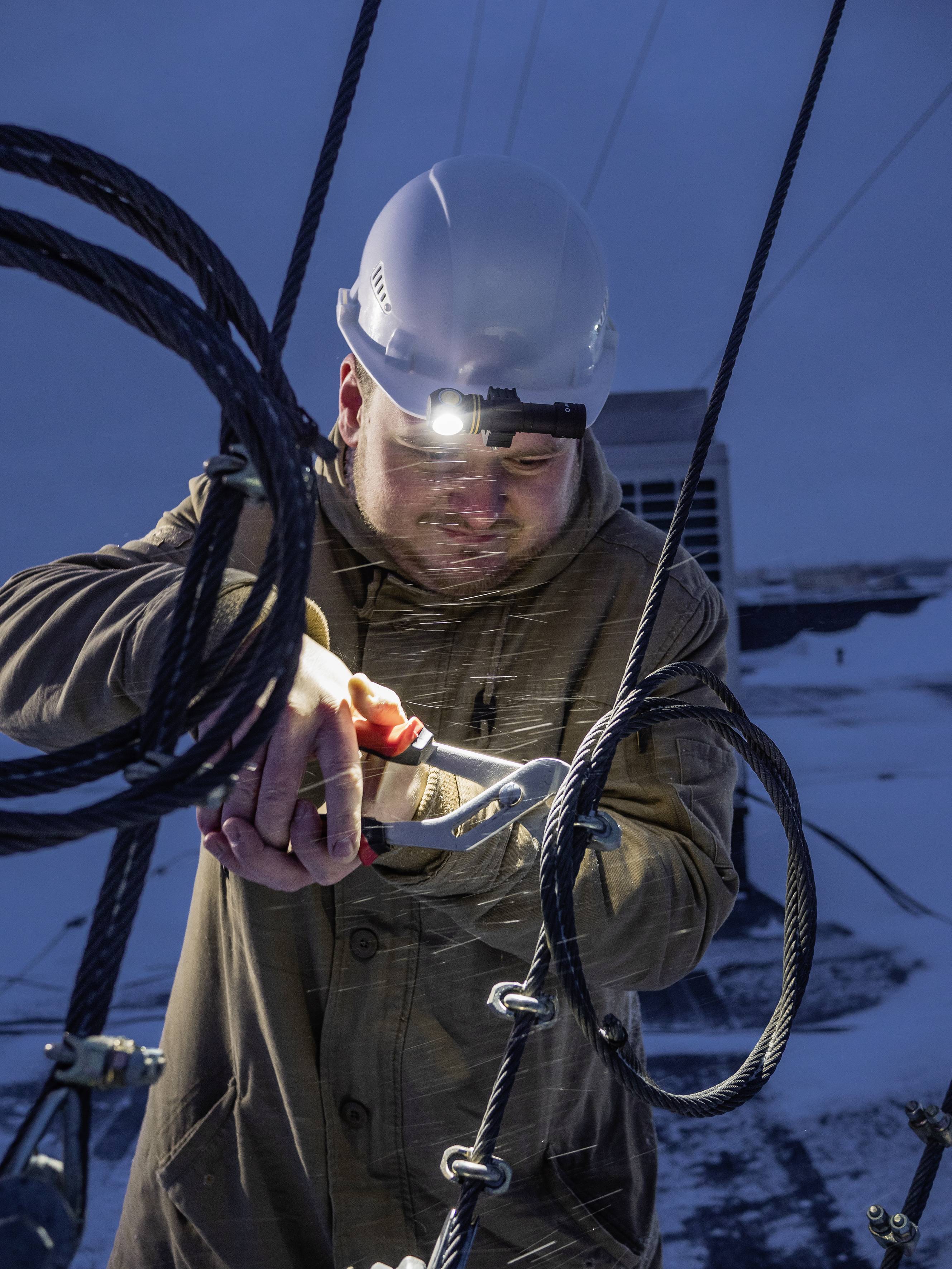 Ein Arbeiter mit Helm und Stirnlampe repariert bei Dämmerung Kabel im Freien. Hintergrund zeigt Schnee und technische Anlagen.