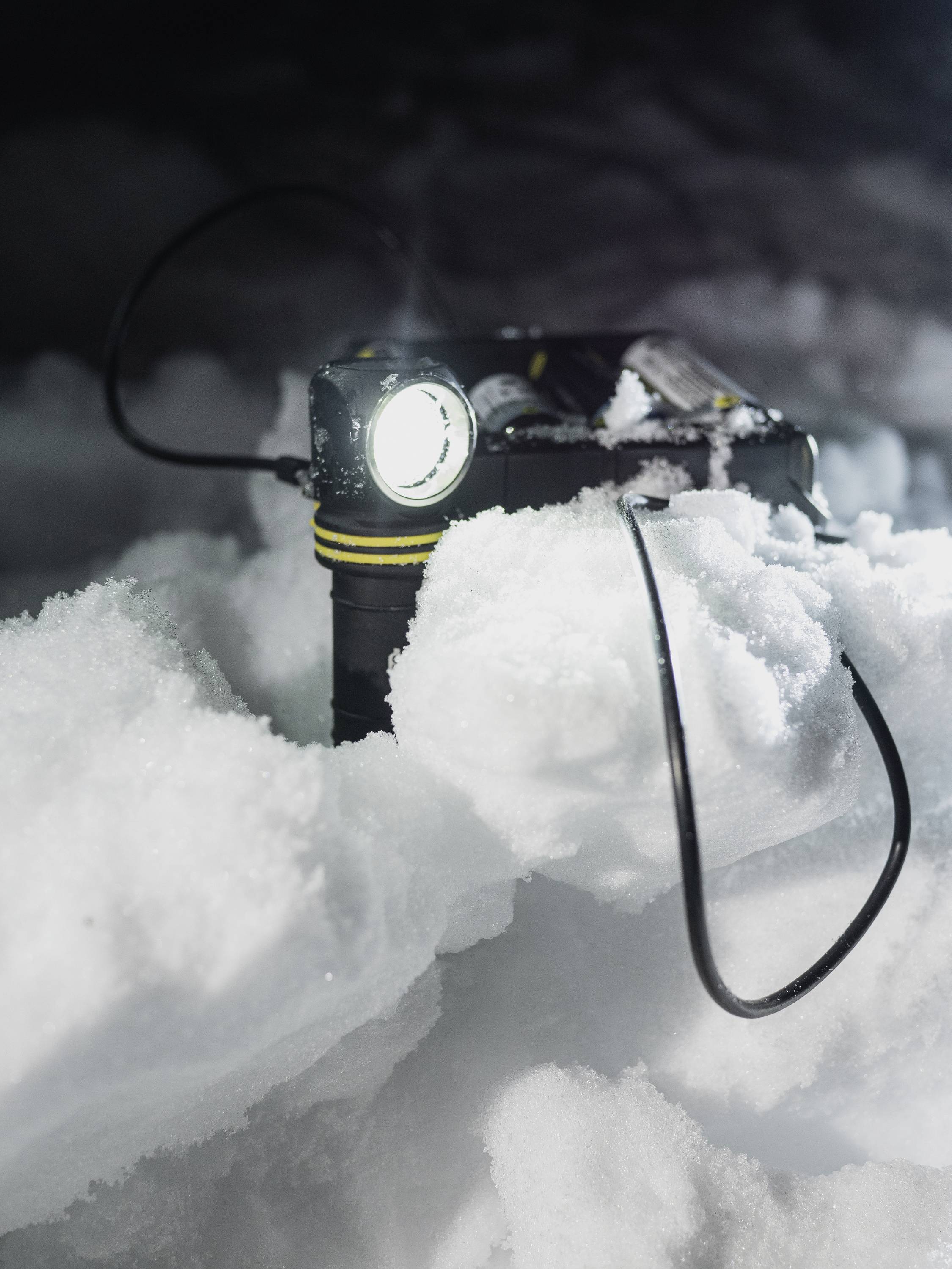 Taschenlampe im Schnee; leuchtet hell in dunkler Umgebung, von Kabel umgeben.