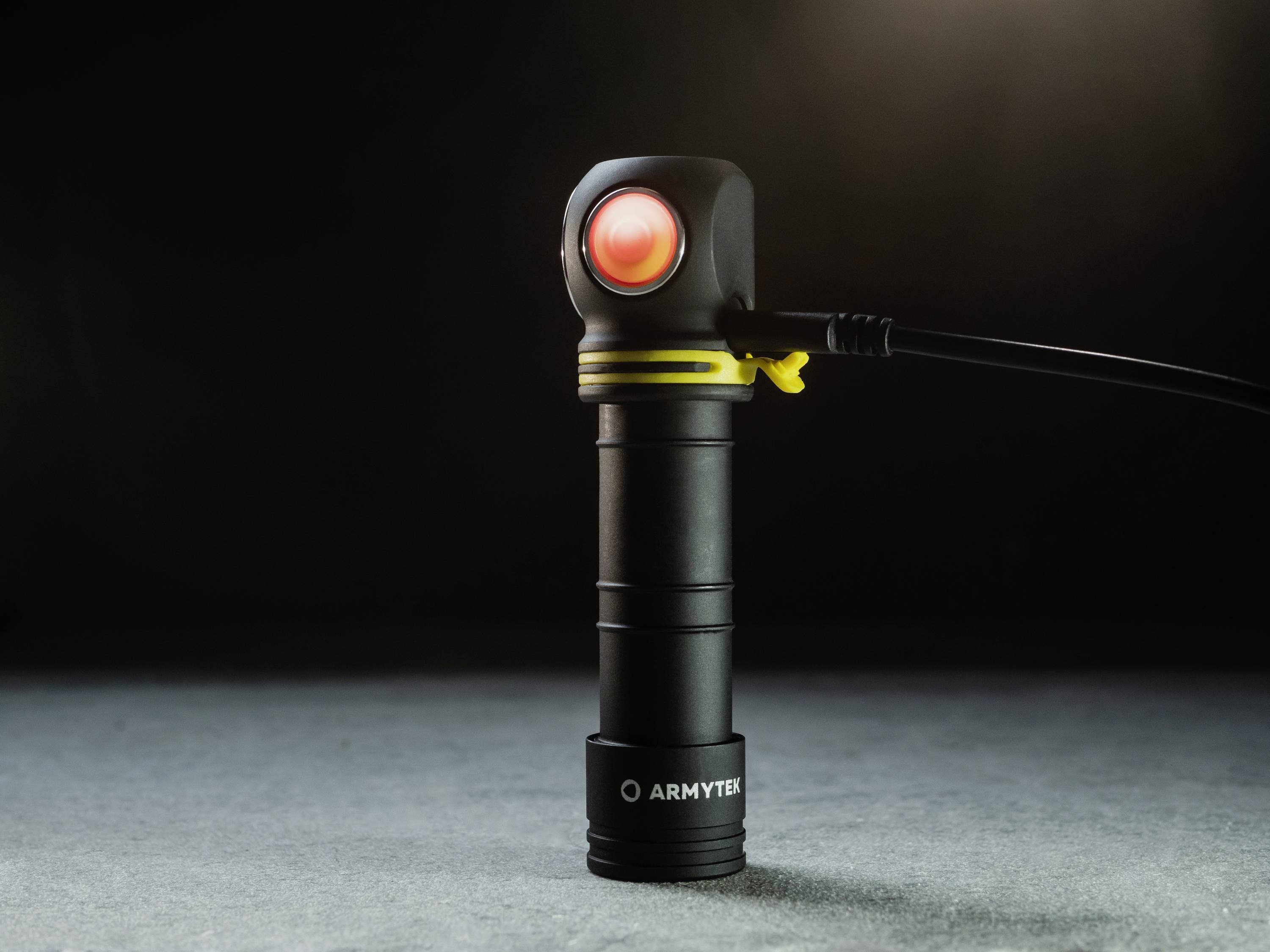 Schwarze Taschenlampe mit rotem Licht leuchtet auf grauer Oberfläche. Ein Kabel ist an der Seite eingesteckt.