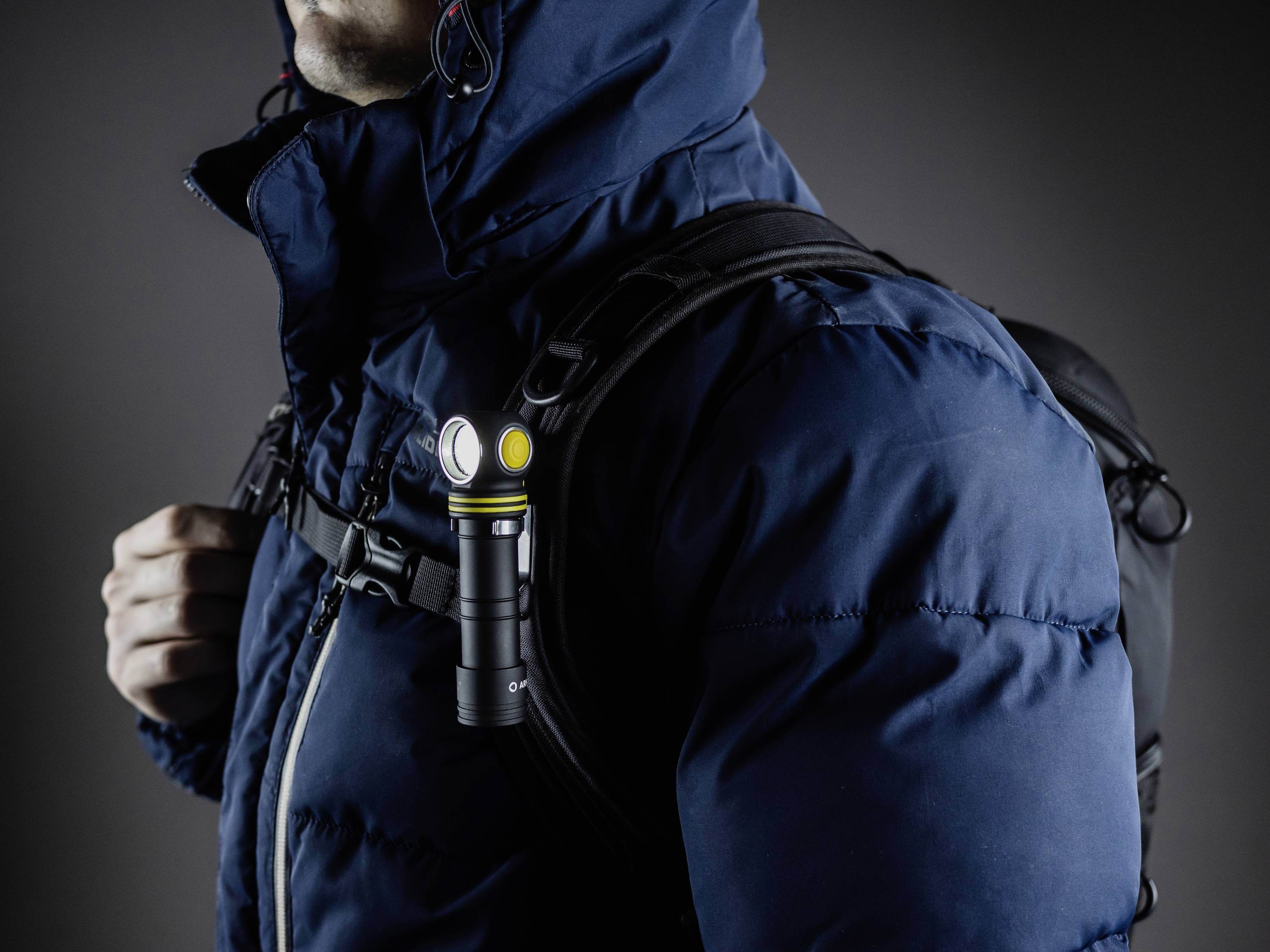Eine Person in einer blauen Winterjacke trägt einen Rucksack. An einem Gurt ist eine kleine, leuchtende Taschenlampe befestigt.