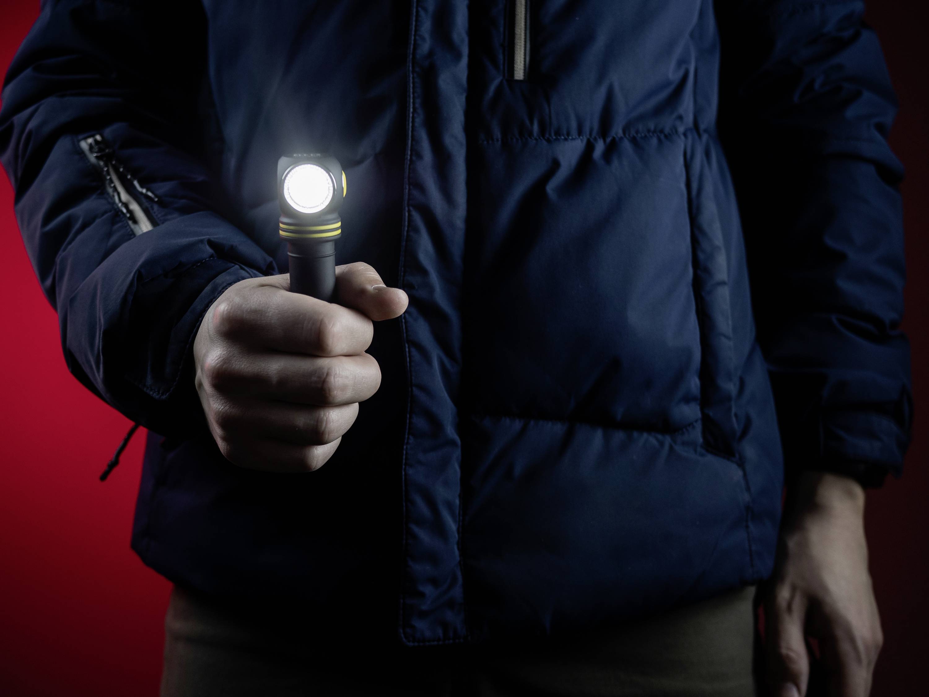 Person in blauer Jacke hält eine eingeschaltete Taschenlampe in der Hand vor einem roten Hintergrund.