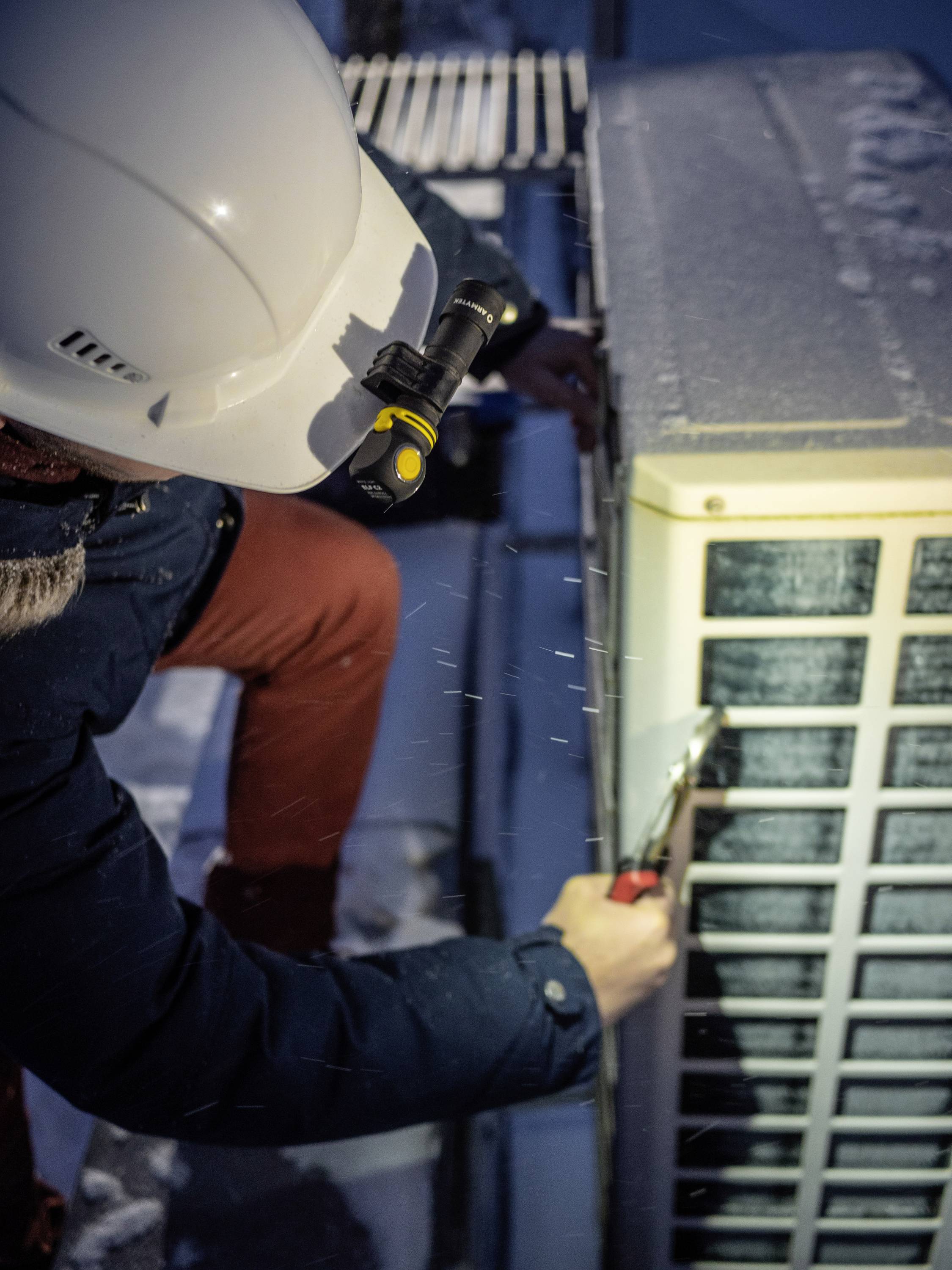 Techniker trägt Helm und arbeitet bei Schnee an einer Klimaanlage. Er benutzt Werkzeug, um das Gerät bei kalten Bedingungen zu warten.