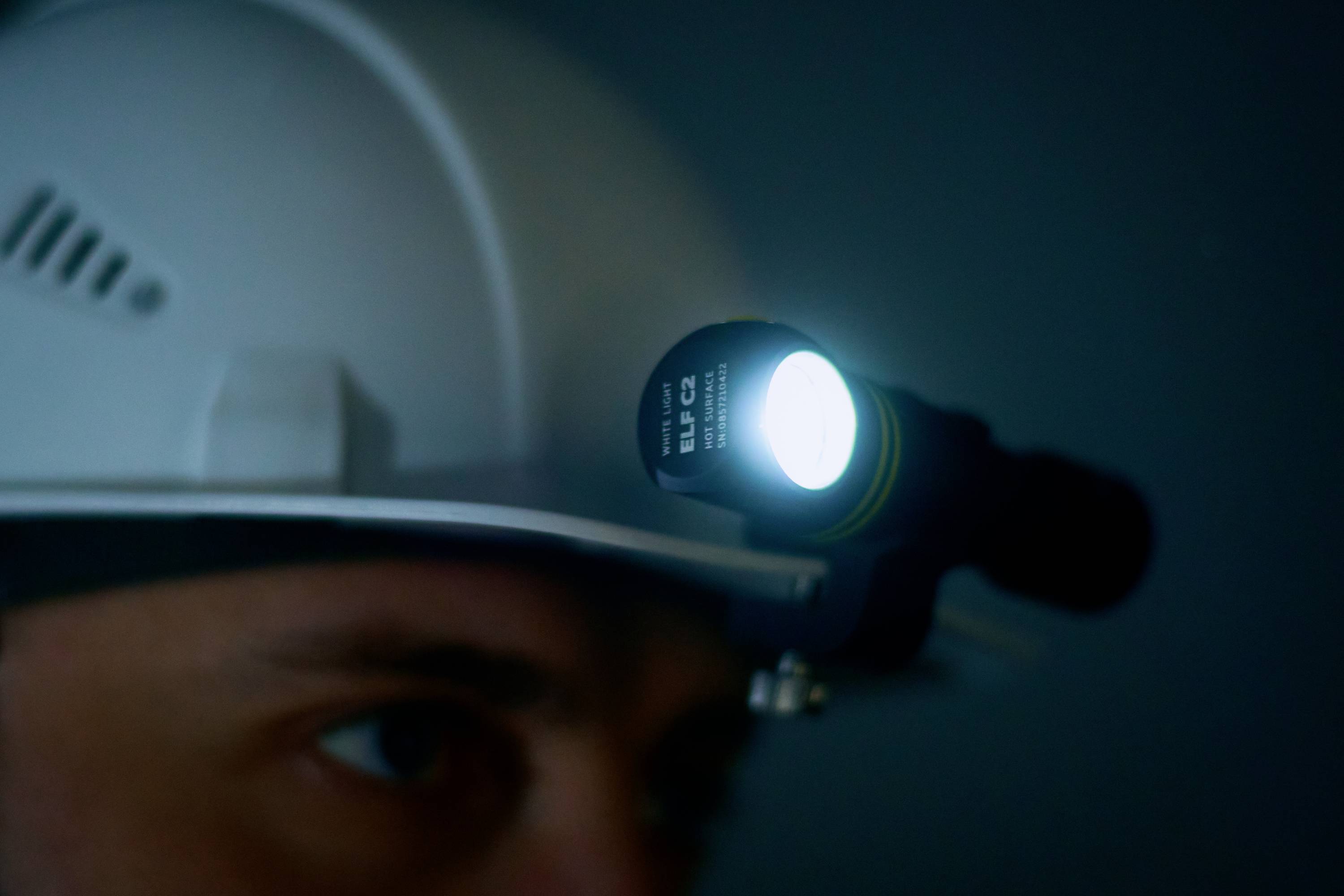 Eine Person trägt einen Schutzhelm mit einer leuchtenden Stirnlampe in einem dunklen Raum, was auf Arbeit in dunkler Umgebung hinweist.