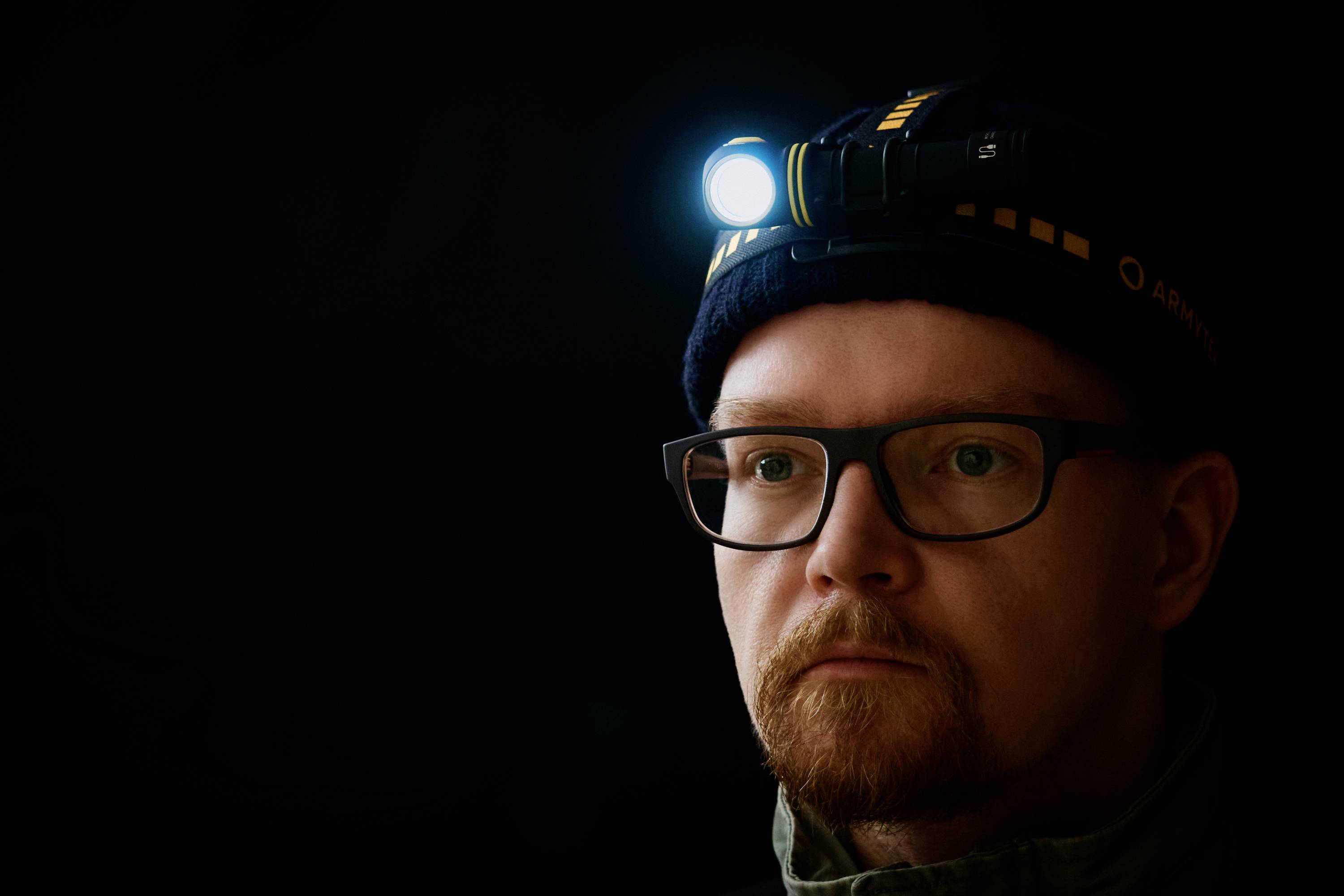 Ein Mann mit Brille trägt eine Stirnlampe, die in einem dunklen Raum leuchtet.