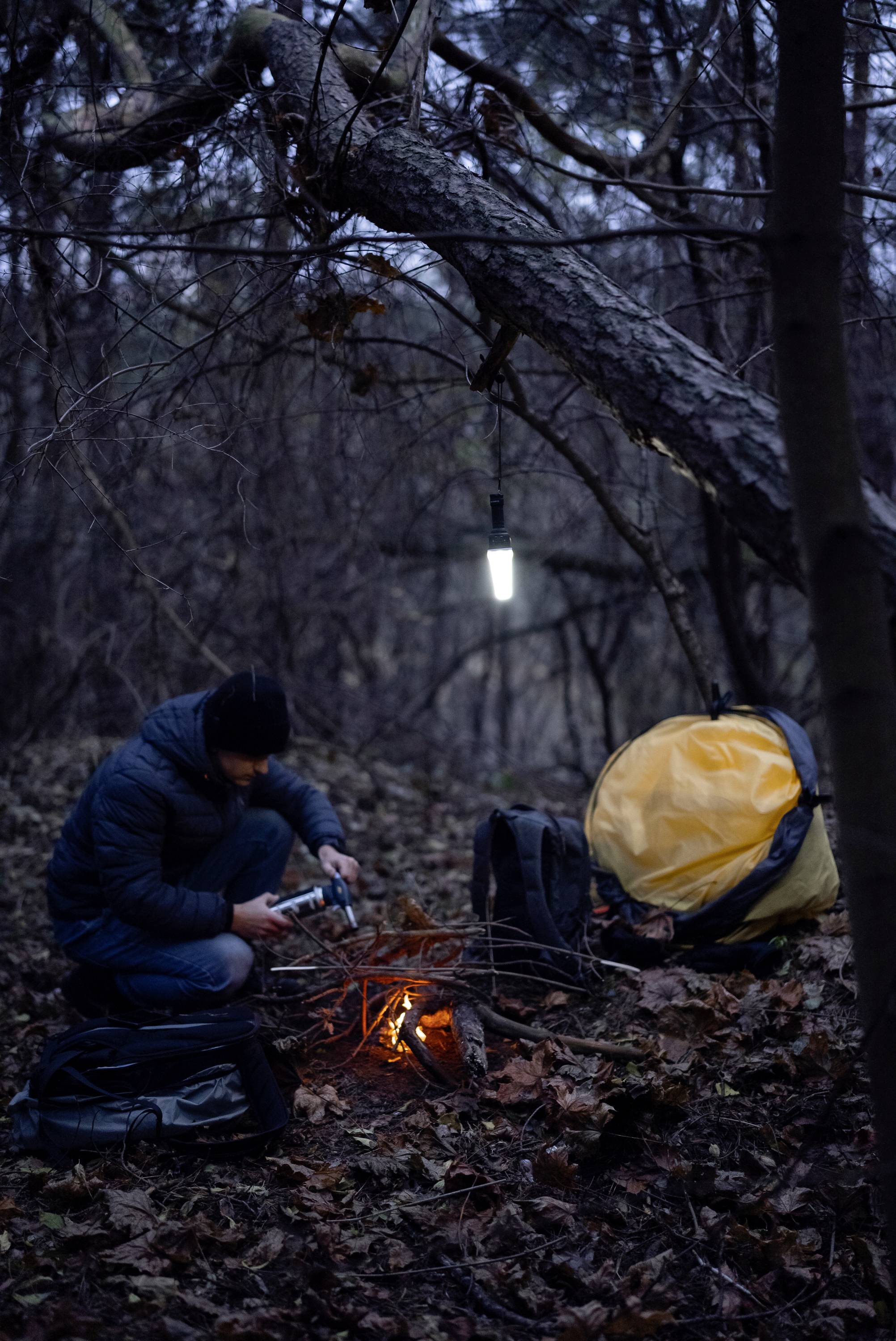Person zündet Lagerfeuer im Wald an, mit Zelt und Taschen in der Nähe, bei Dämmerung.