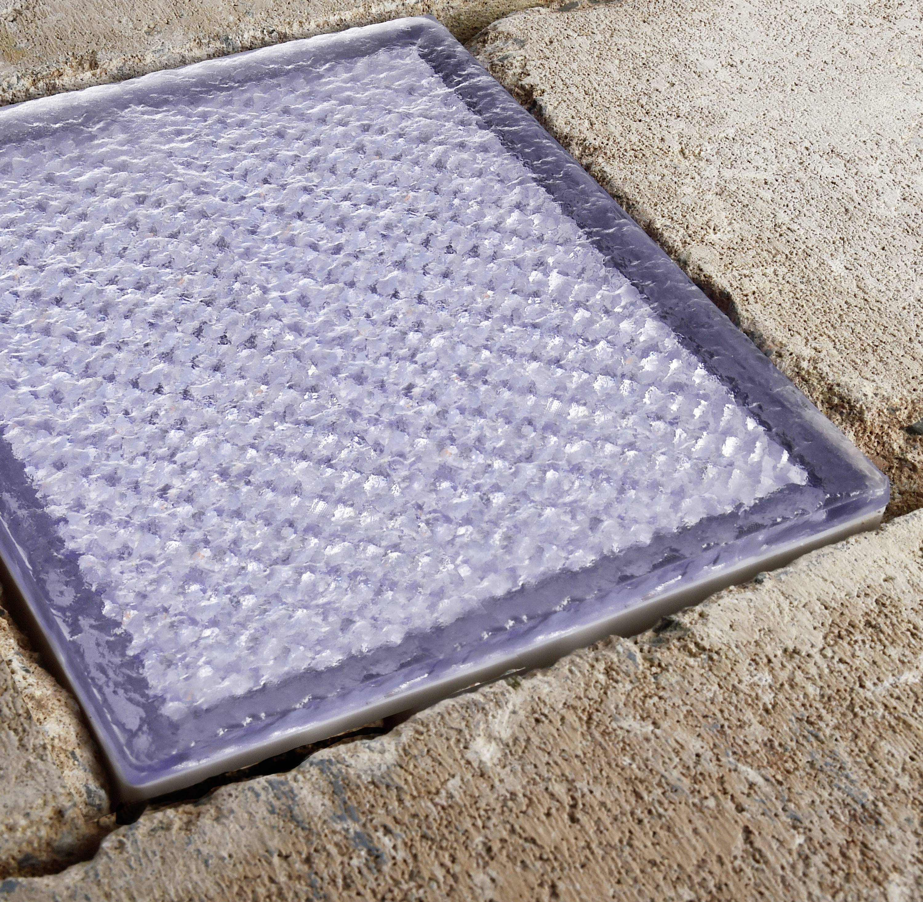 Eine quadratische, hellblaue Glasplatte mit strukturierter Oberfläche auf einer sandfarbenen, gepflasterten Steinfläche.