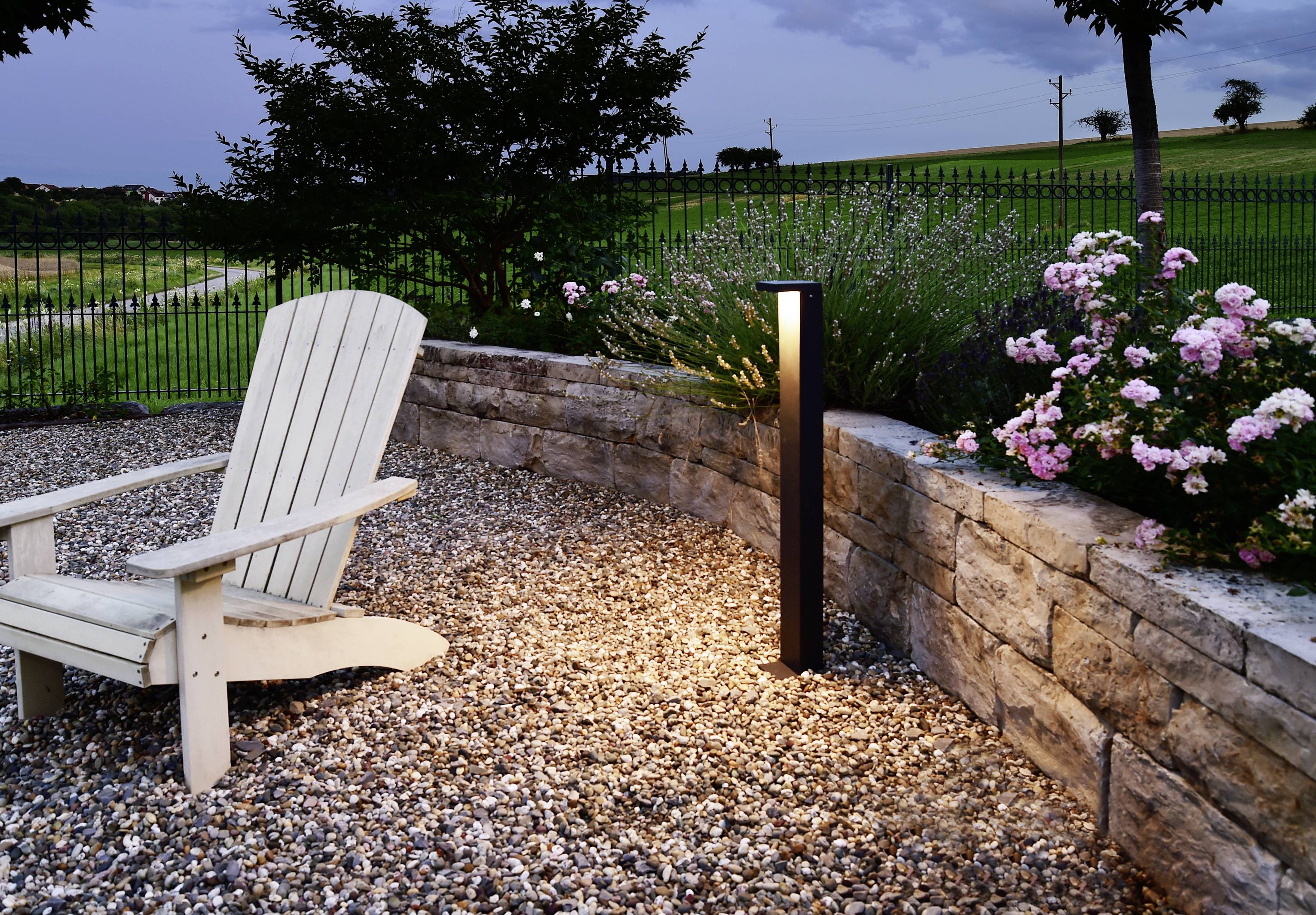 Ein Garten mit einer Steinmauer, beleuchtet von einer Standleuchte. Eine weiße Holzstuhl steht auf einem Kiesweg, umgeben von Blumen.