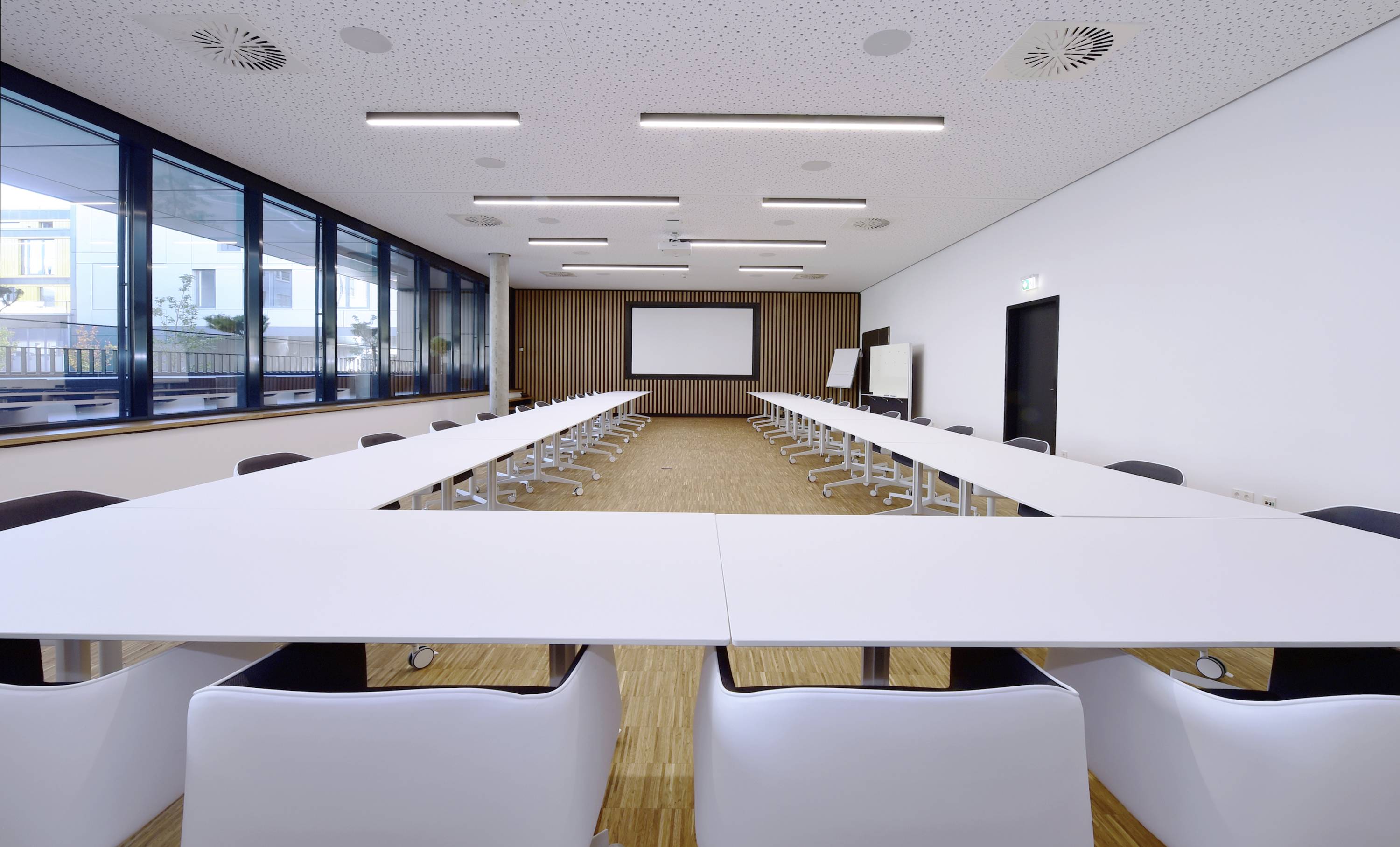 Ein moderner Konferenzraum mit U-förmiger Tischanordnung, weißen Stühlen, großen Fenstern und einem Bildschirm vorne an der Wand.