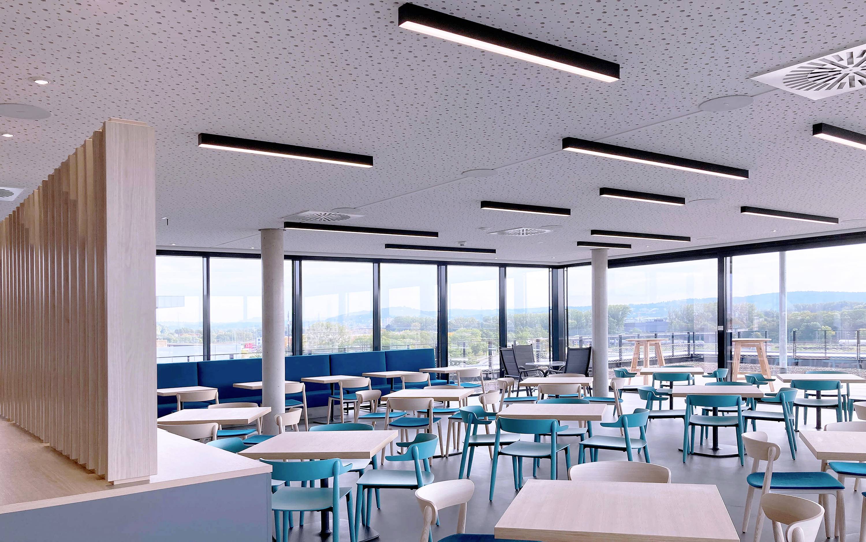 Ein modernes, leeres Café mit Holzmöbeln, blauen Stühlen und großen Fenstern, die einen Blick auf eine Außenlandschaft bieten.