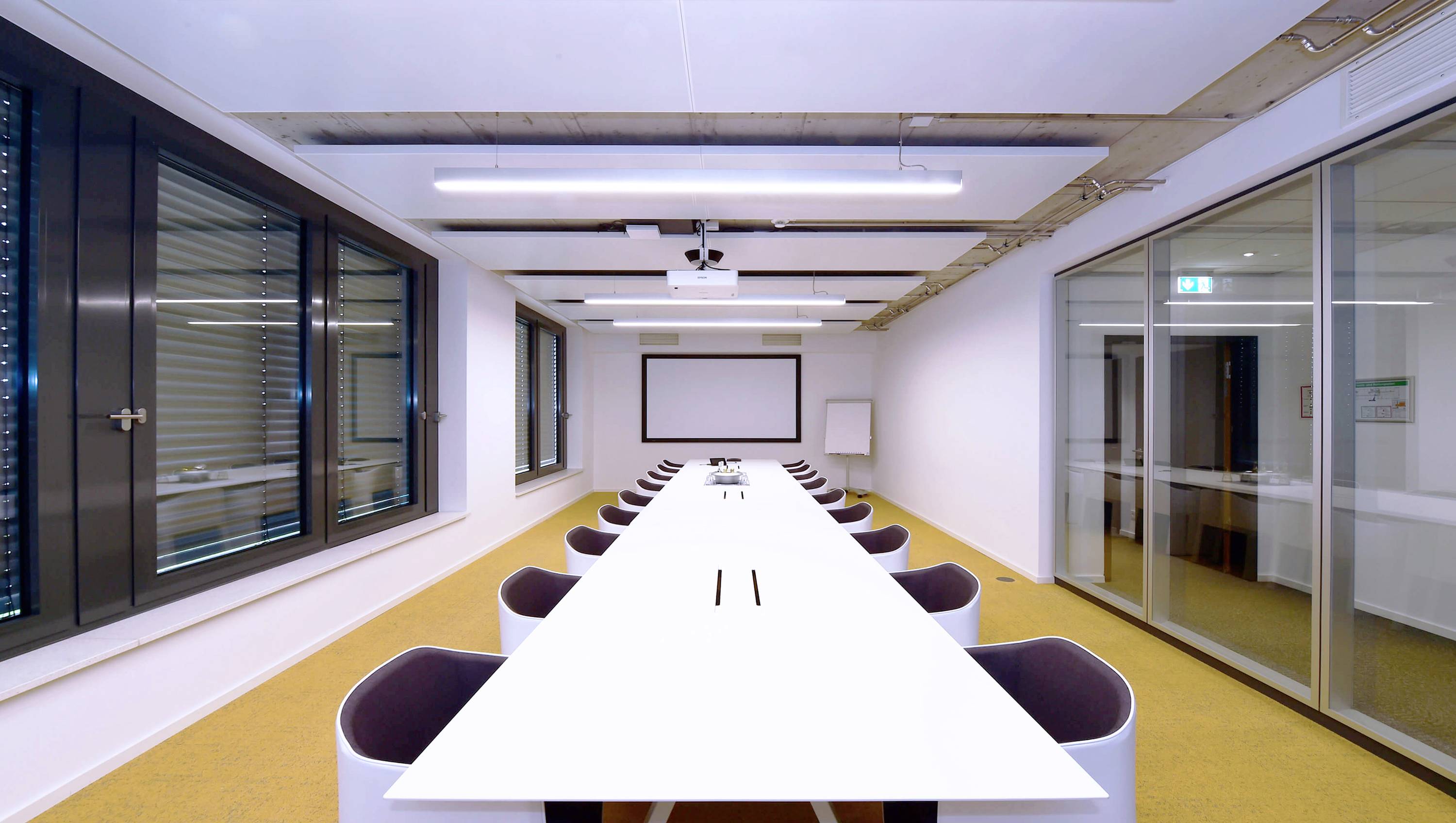 Ein moderner Besprechungsraum mit langem weißem Konferenztisch, weißen Stühlen, einer Leinwand an der Wand und Glasfenstern auf der linken Seite.