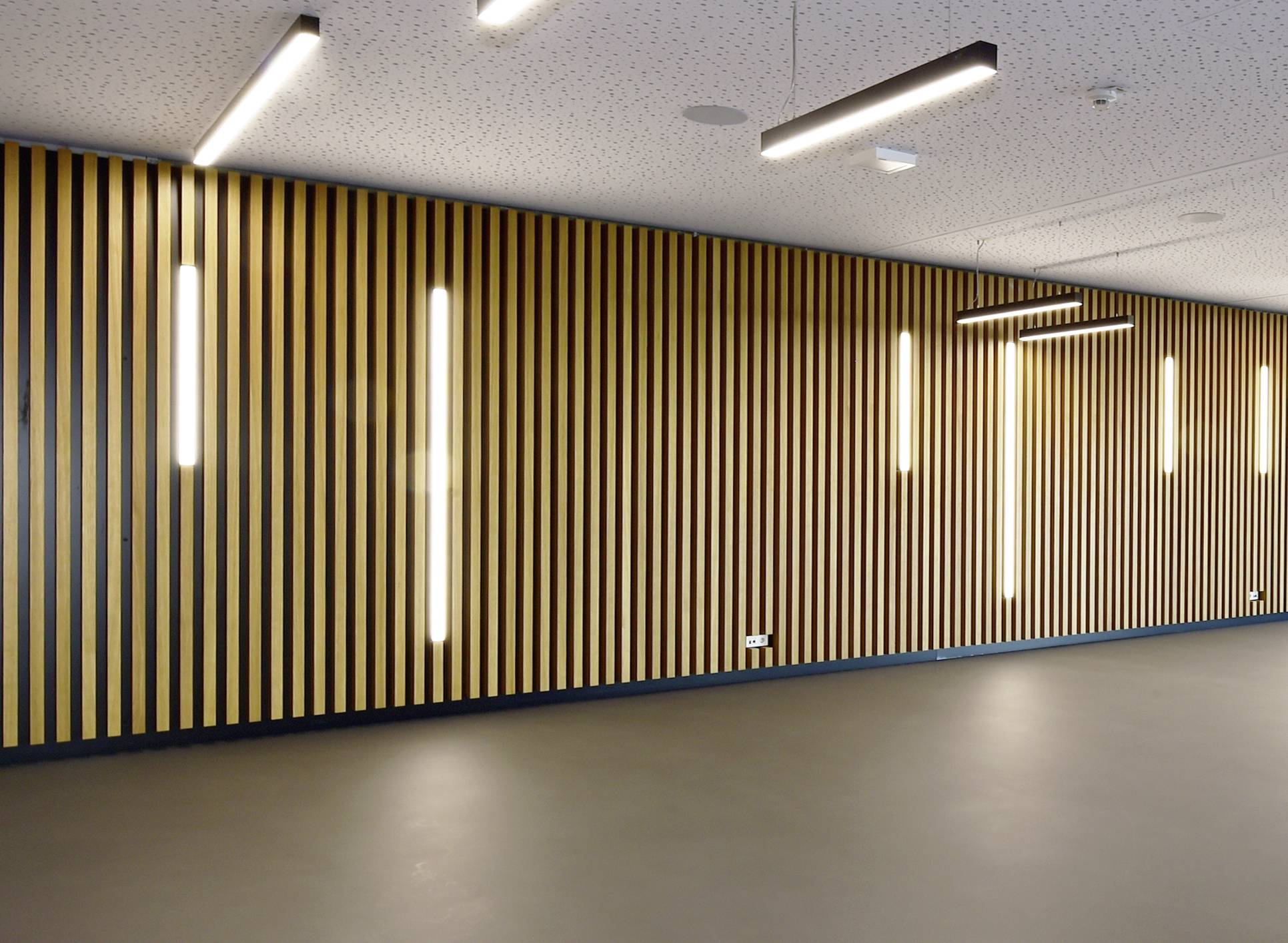 Ein Raum mit einer Wand aus vertikalen Holzlatten und mehreren modernen, länglichen Deckenleuchten. Der Boden ist glatt und grau.