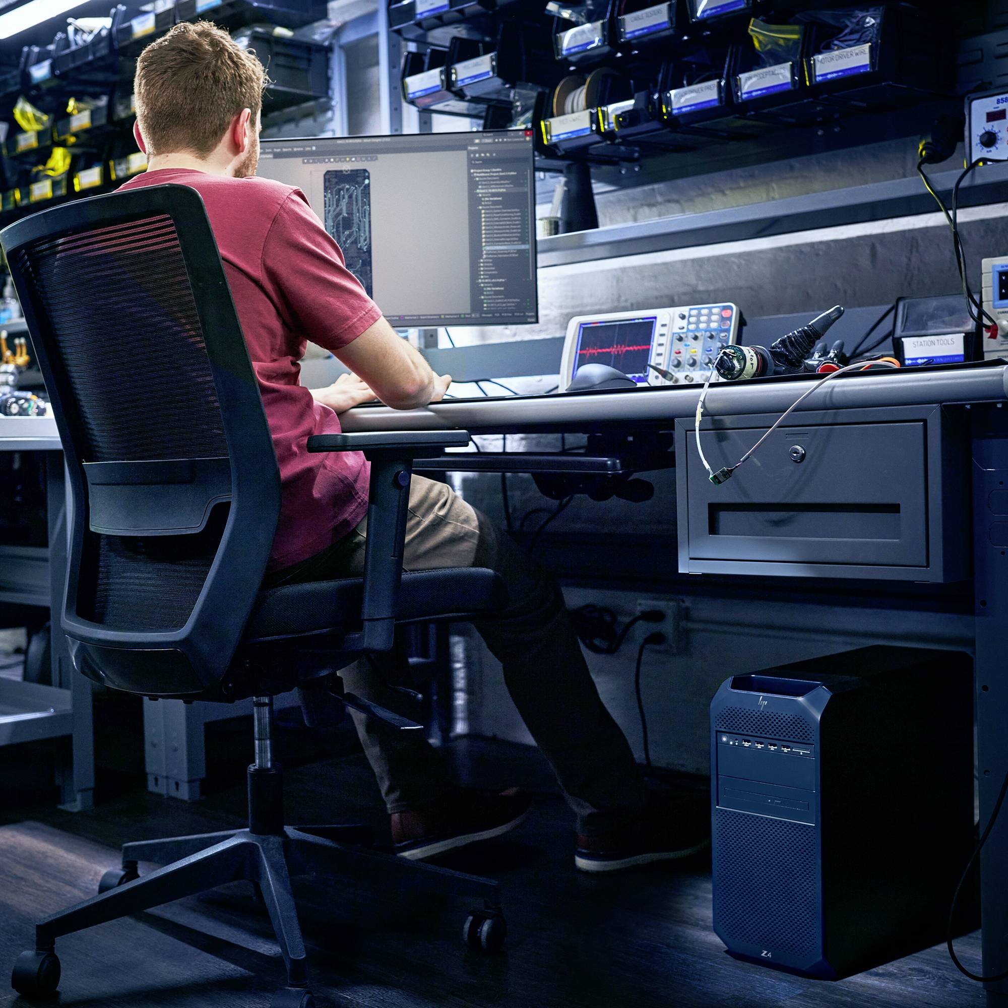 Ein Mann sitzt an einem Schreibtisch und schaut konzentriert auf Computerbildschirme mit Code. Auf dem Tisch sind verschiedene Technikgeräte.